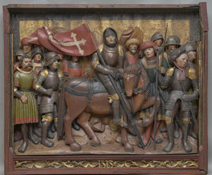 A multi-colored wood sculpture in a rectangular red frame with a group of people in military armor surrounding one horse-mounted figure in military armor holding a flag on a pole.
