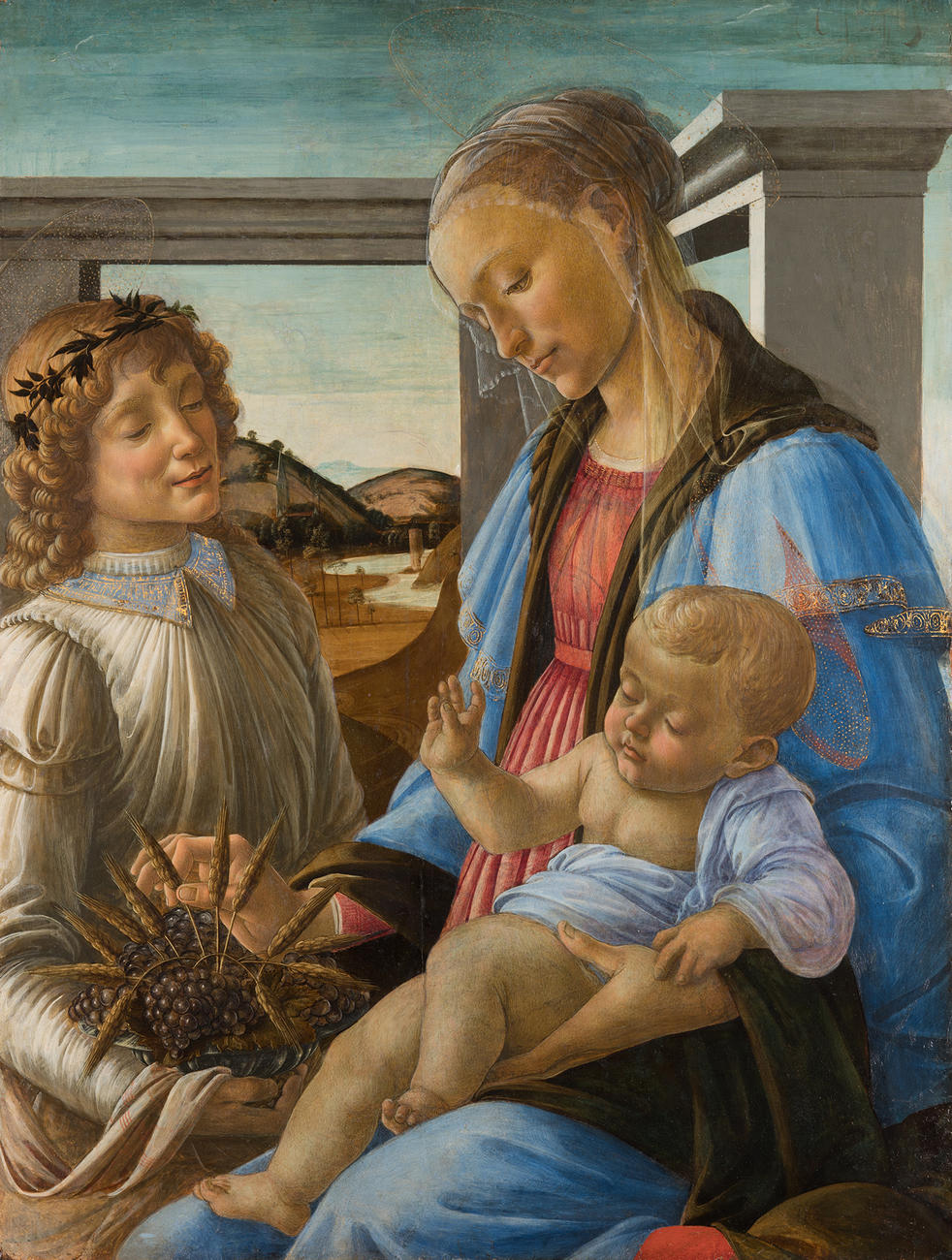 Boticelli’s painting Virgin and Child with an angel before the 2023 conservation treatment. A woman, the Virgin Mary, with a halo, veil, and wearing a blue robe holding an infant, the baby Jesus, in her lap. Standing to her left is a man, an angel, with a halo and wearing a white robe. He holds a pomegranate with sprigs of wheat. Behind them are white columns and a landscape.