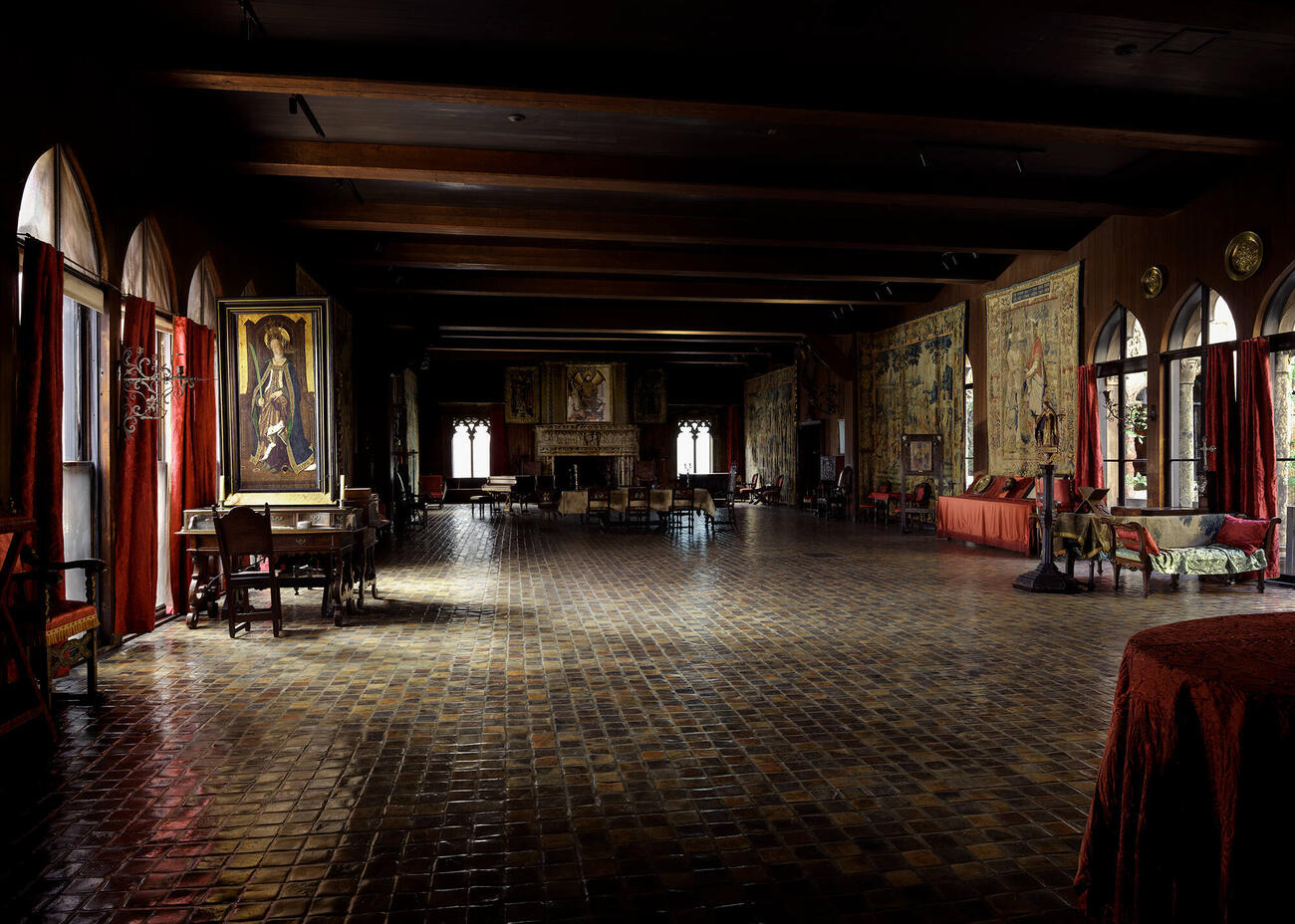 South-facing view of the spacious Tapestry Room gallery from 2011, with tiled floor, wooden beamed ceiling, large tapestries displayed on the walls as well as prominent paintings and a large carved stone fireplace.