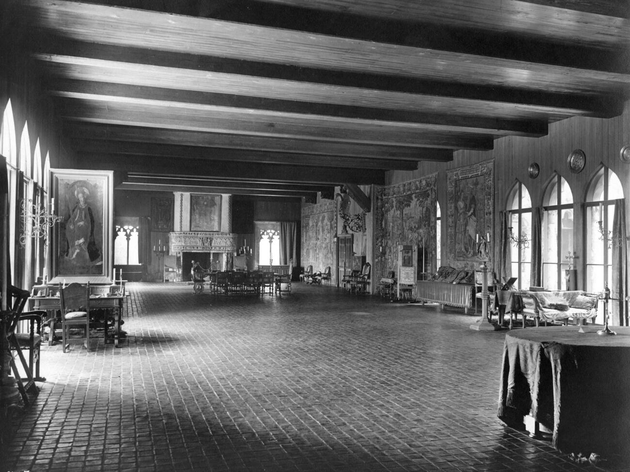Historic black-and-white south-facing view of the spacious Tapestry Room gallery from 1926, with tiled floor, wooden beamed ceiling, large tapestries displayed on the walls as well as prominent paintings and a large carved stone fireplace.