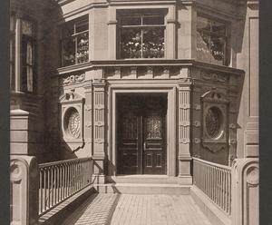 Exterior of Isabella Stewart Gardner’s Neo-Classical style brownstone home at 152 Beacon Street. A railed walkway leads to the large double door with wrought iron windows. The entrance is flanked by fluted pilasters punctuated by reliefs of a single flower. Above the front door on the second story, the three bay  windows are filled with displays of flowers. 