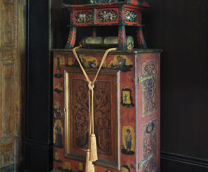 A wooden cabinet painted red with pastoral figures is the base of a pyramidal installation in the Early Italian Room of the Isabella Stewart Gardner Museum. On top of the cabinet is a Japanese hanging amulet with a long yellow cord.  Above it, a black ceramic figure of Chinese poet Li Po sits on top of a Japanese red and black lacquer writing desk.