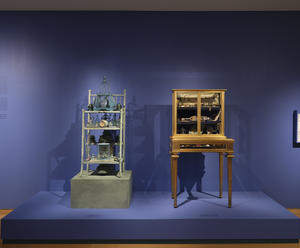 An assemblage by artist Betye Saar of a seafoam green set of three shelves with attached clocks, birdcages, and glass containers and a gilt vitrine curated by Isabella Stewart Gardner displaying many small objects. 