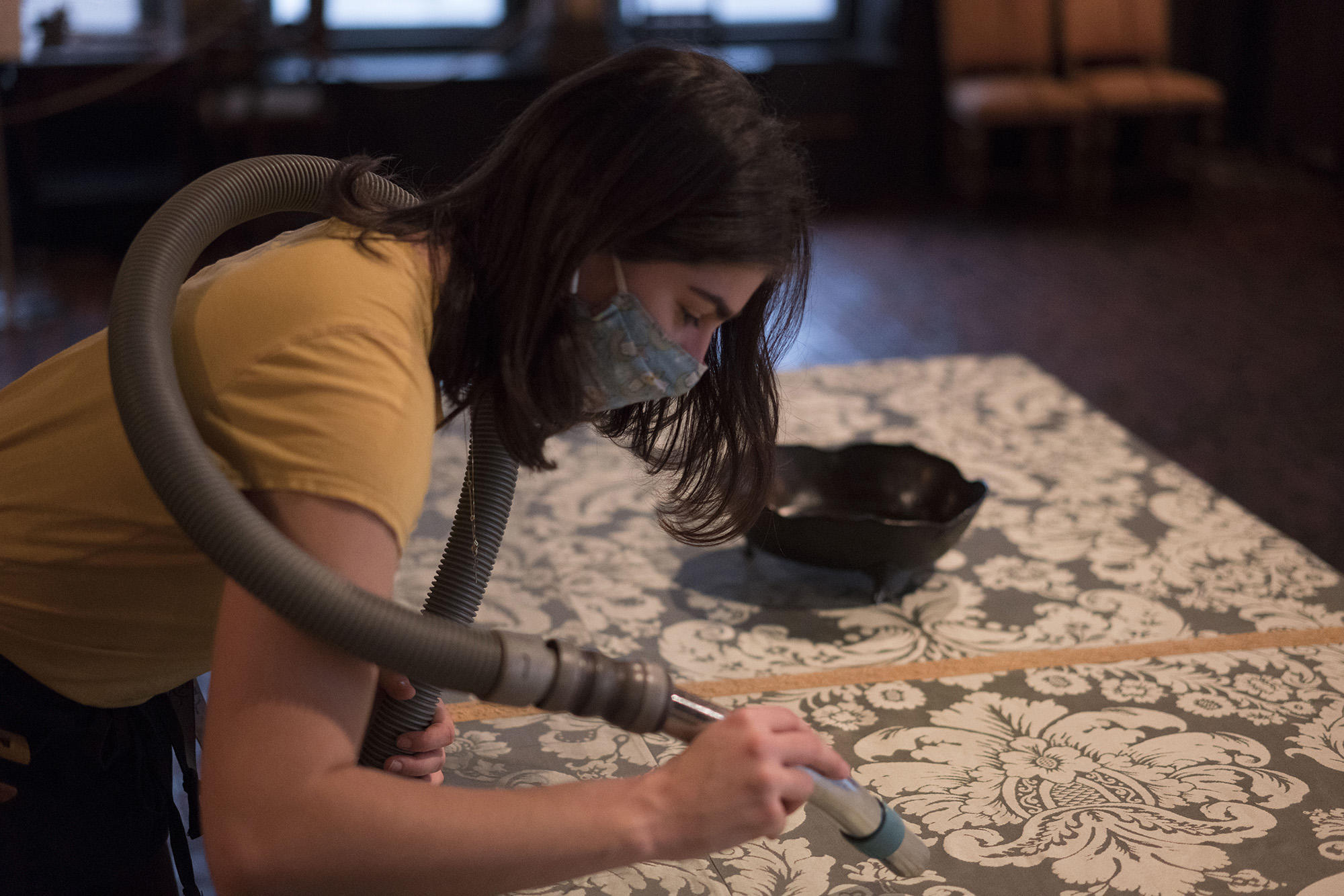 Vanessa Escamilla hovers over a textile table covering to remove dust, 2021