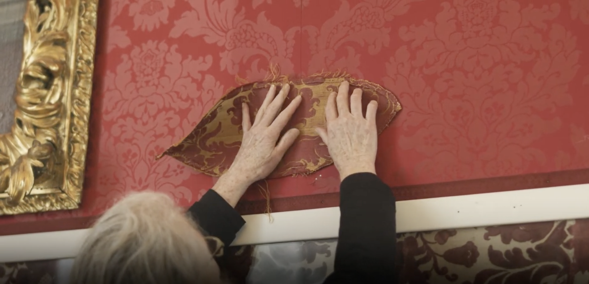 A person holding up a scrap of tapestry on a wall.