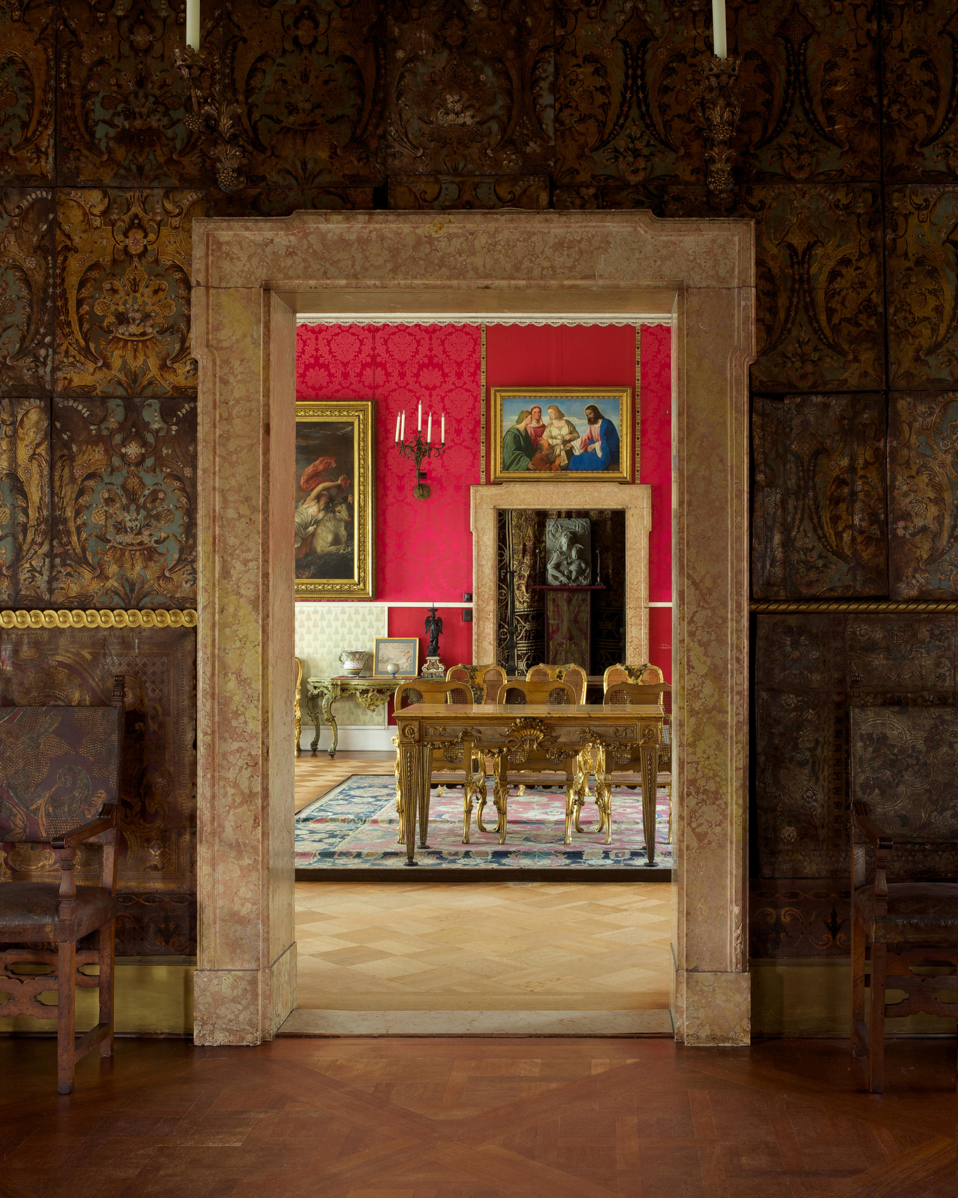 A room covered in brown tapestries with a doorway entering into a red room with paintings. 