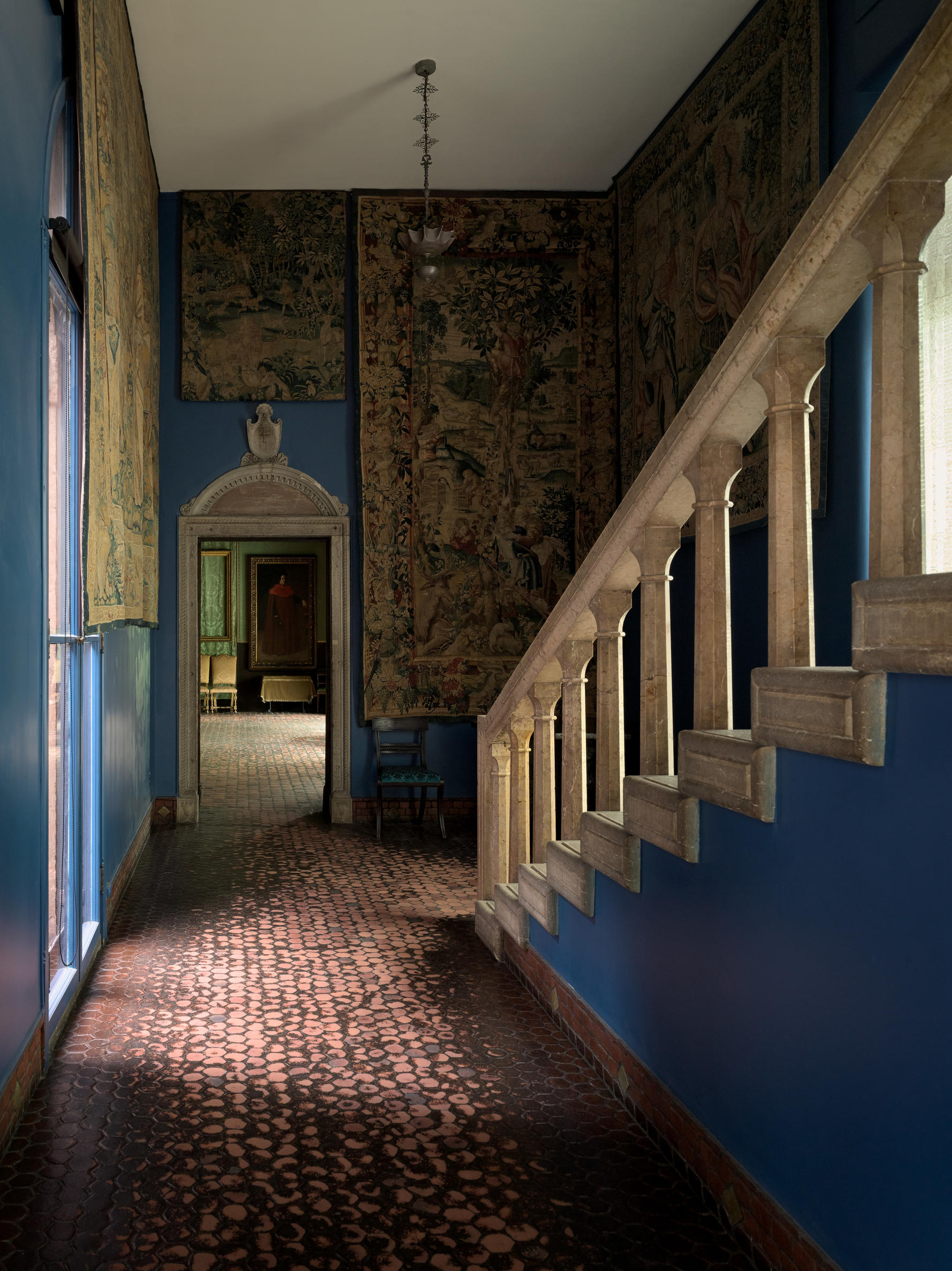 A hallway with a decorated dark red rug and a painted blue staircase on the right. There is a doorway at the end of the hallway.