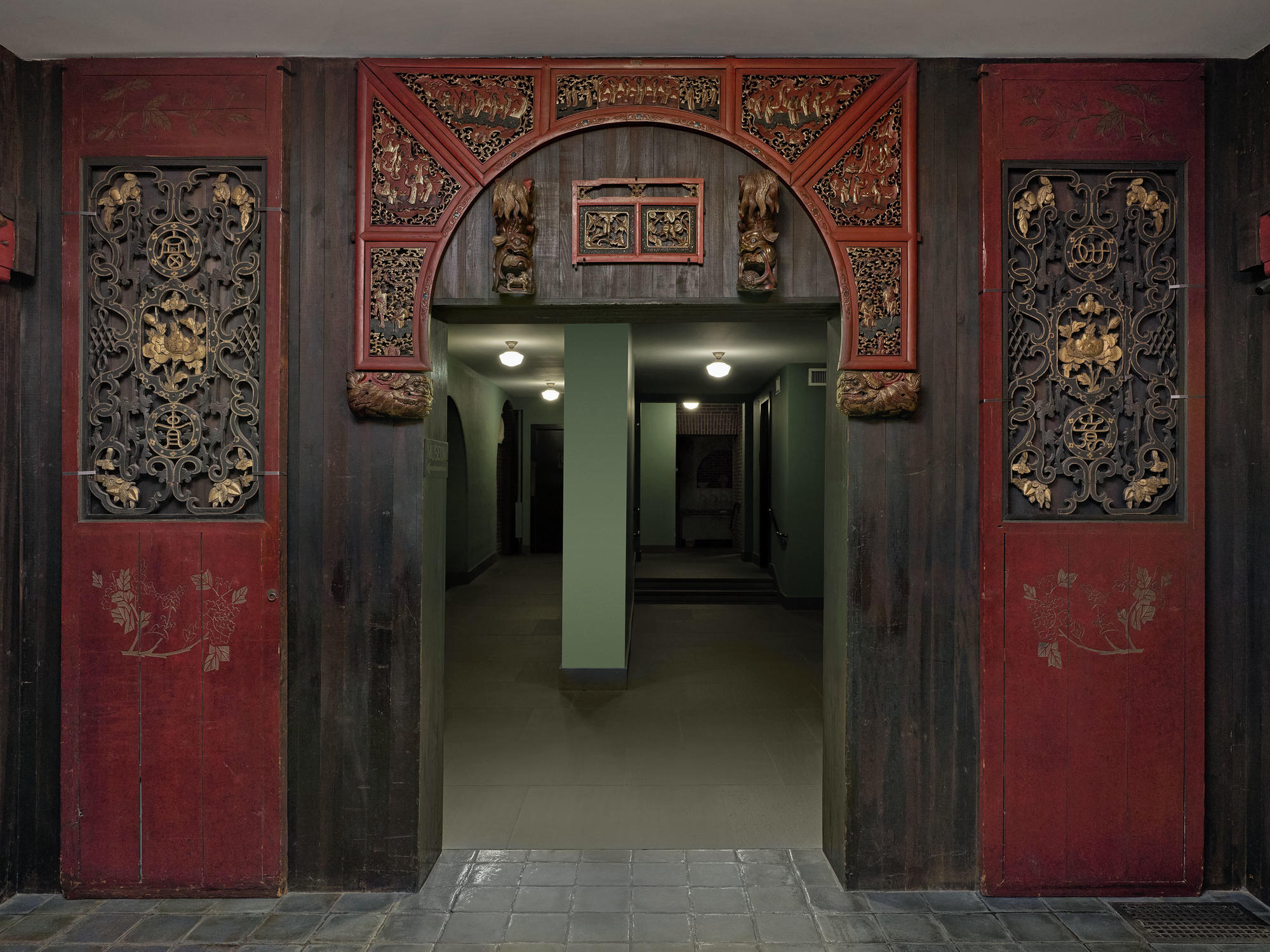 A dark red room with panels on the walls with a doorway leading into a green room with a column in the middle of that room. 