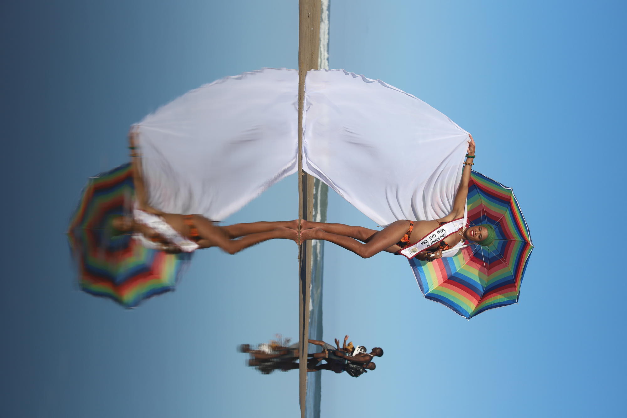 A photo of a person with a white cape, sash and rainbow umbrella flag on the beach.