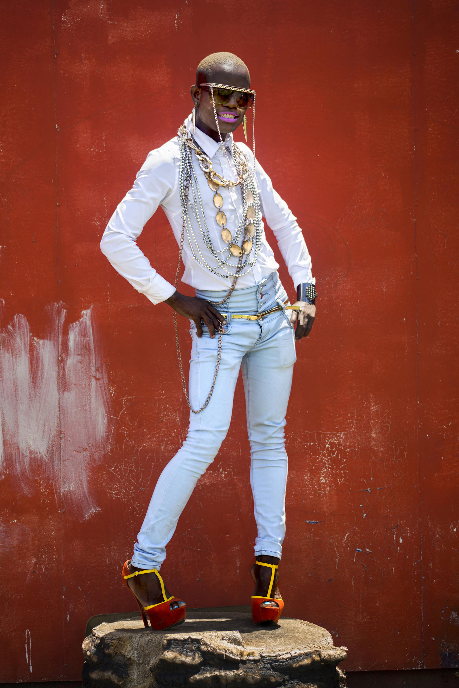 A photo of a person with long necklaces, a white shirt, sunglasses, skinny jeans and red heels stands in front of a red wall.