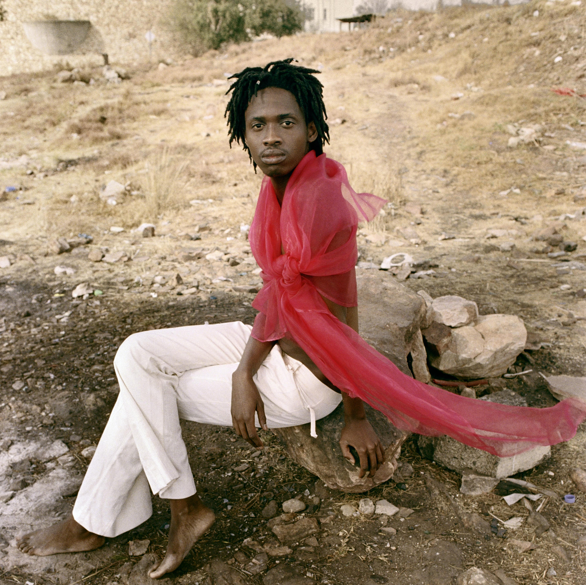 A photo of a person wearing a red sash and white pants sitting on dirt and rocks.