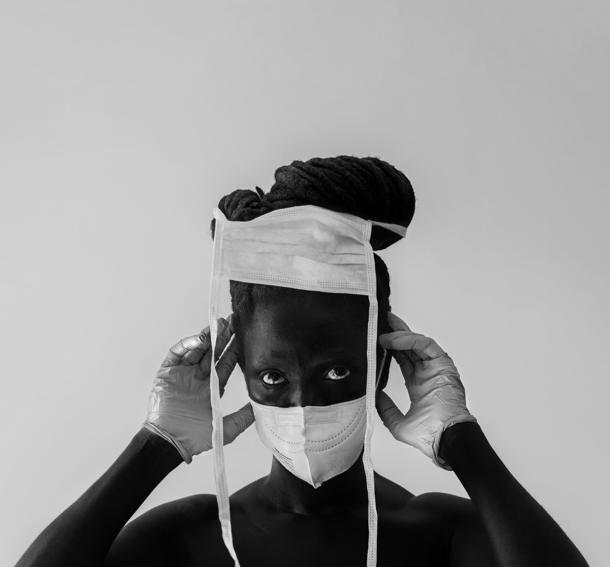 A black and white image of a person with a surgical mask on, gloves and a mask in their hair.