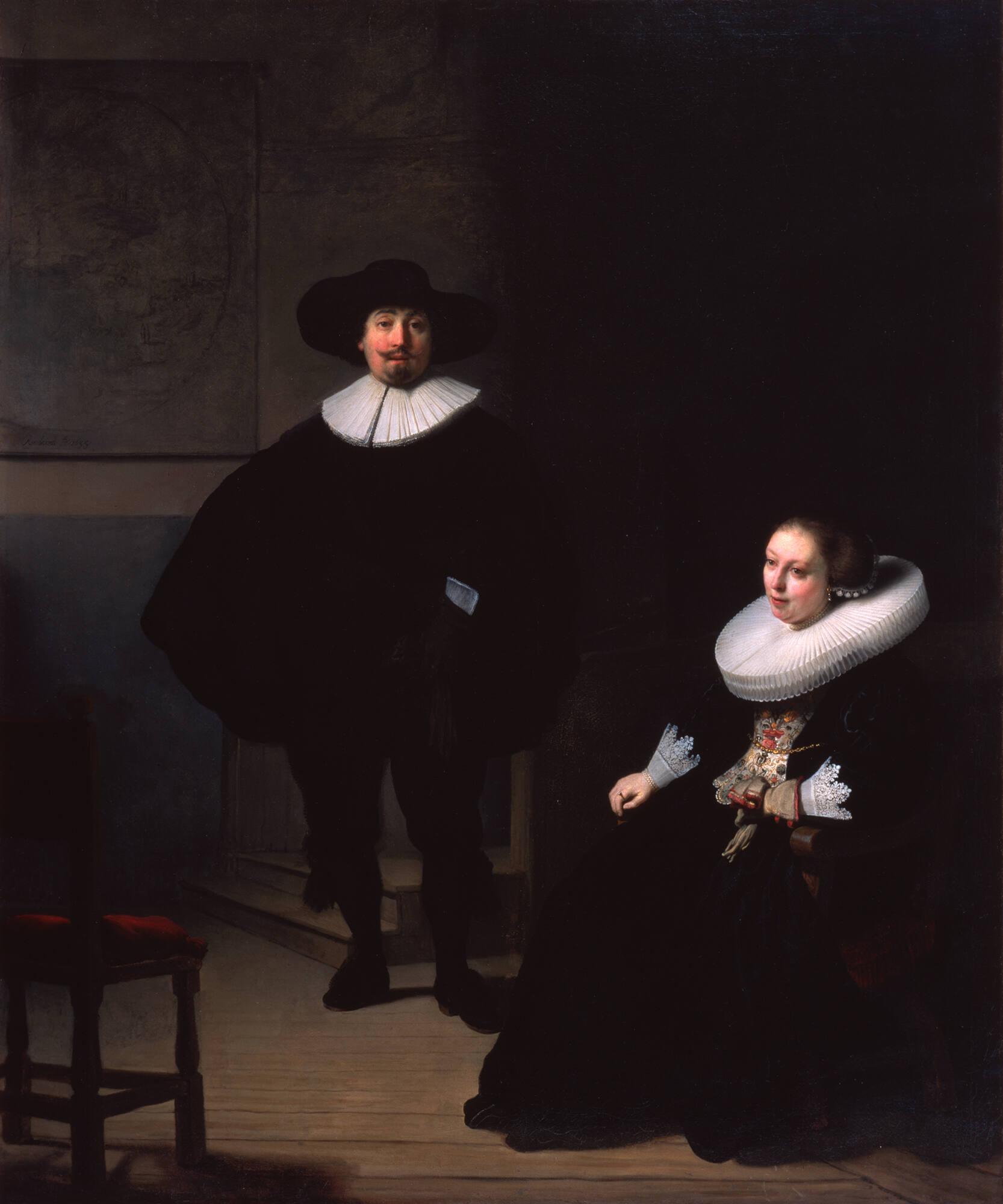 A man standing in a dark room with a hat and a woman sitting next to him.