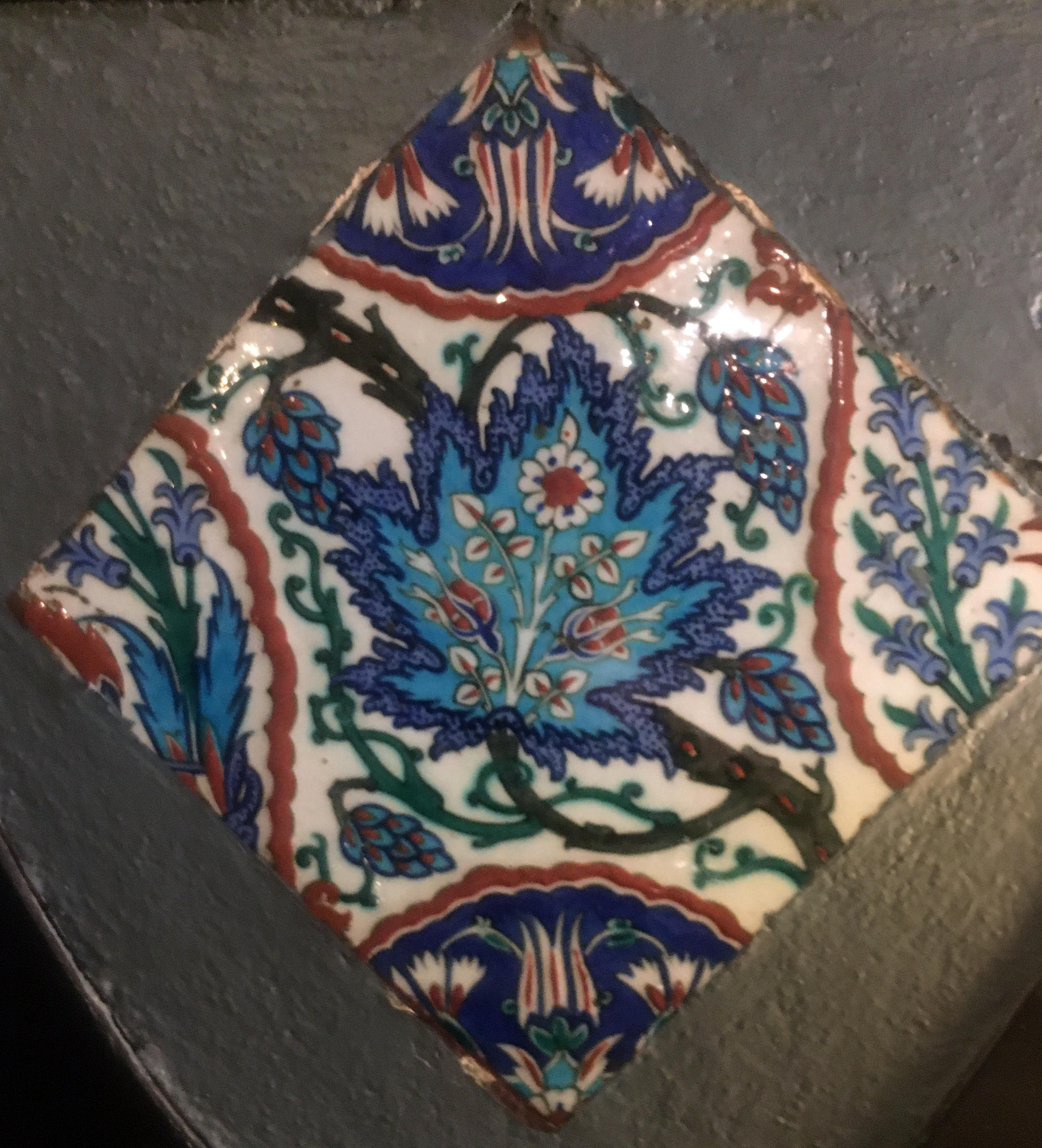 A square blue, white, red, and green ceramic tile with a central stylized grape leaf, nearly identical to the Gardner’s tile.