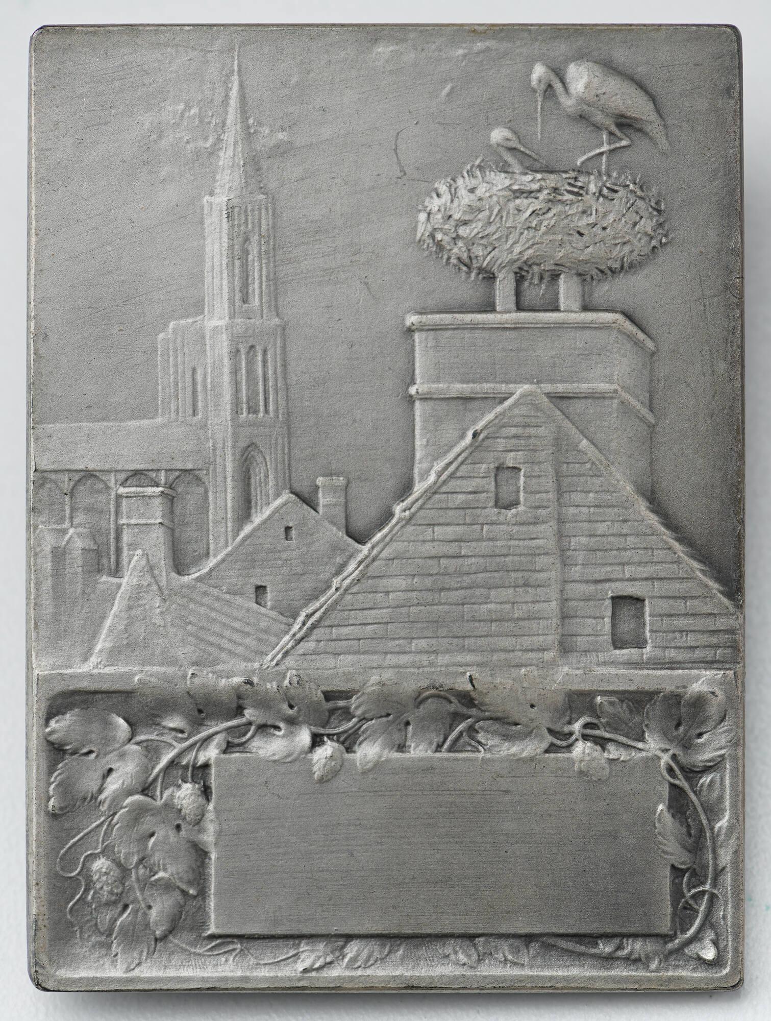 Silver plaquette with a steeple, rooftops, and a stork's nest on top of a chimney in high relief.