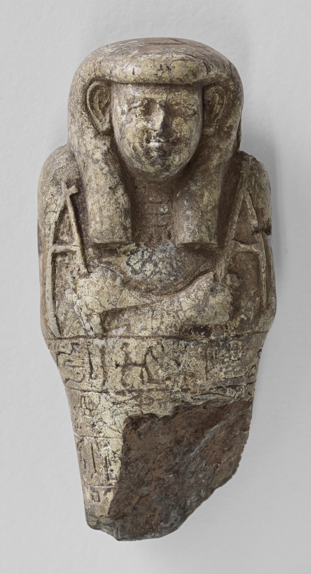 Top half of an Egyptian statuette fragment wearing a ceremonial headdress with arms crossed holding a slender object in each hand.