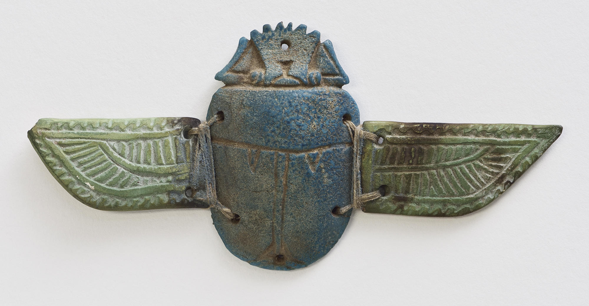 Carved scarab beetle figure in shades of blue with a carved wing attached on either side with twine.