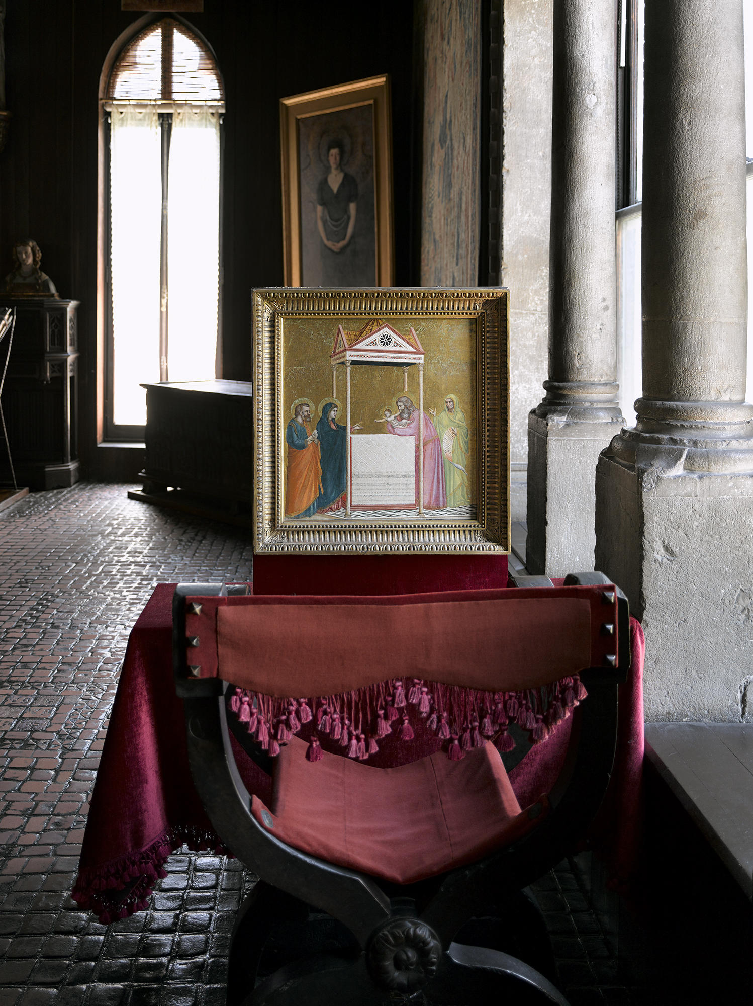  A small Italian Renaissance painting with a gold background on an easel in front of a chair upholstered in red velvet.