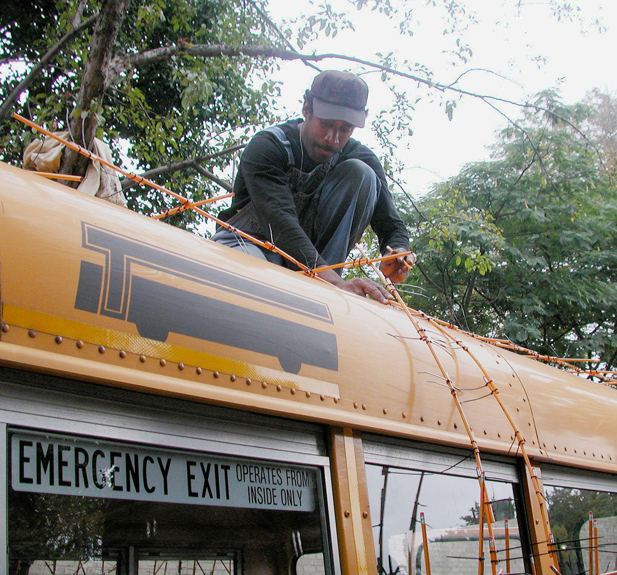 Artist Nari Ward on top of a yellow school bus  constructing a bramble of pencils and zipties.