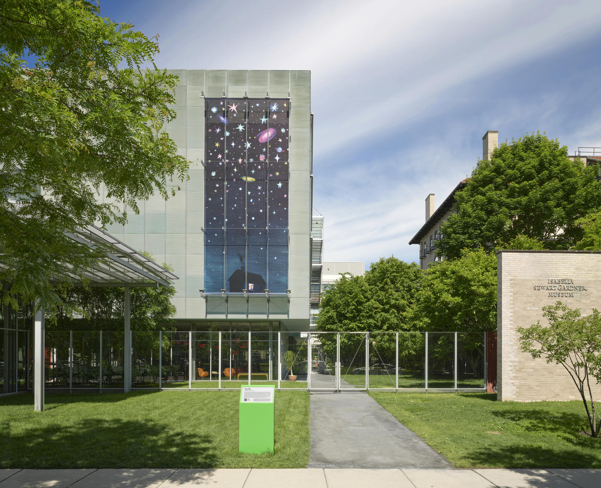 Exterior of the Isabella Stewart Gardner Museum showing a large-scale, 36-foot by 16-foot artwork installed vertically on the outside of the Museum’s New Wing. The artwork depicts a nighttime scene of a house at the bottom with multi-colored stars and galaxies against a dark blue sky above it. 