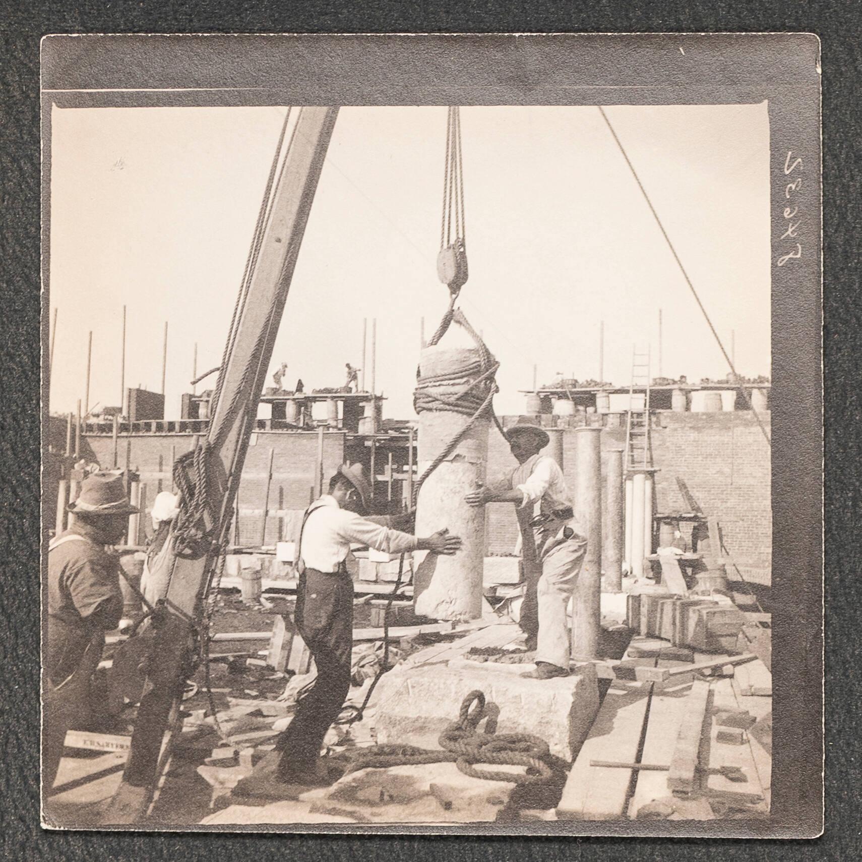 A black and white photograph showing three men on the Gardner Museum construction site with two of the men at the center holding a pillar that is suspended in the air with rope on a crane.