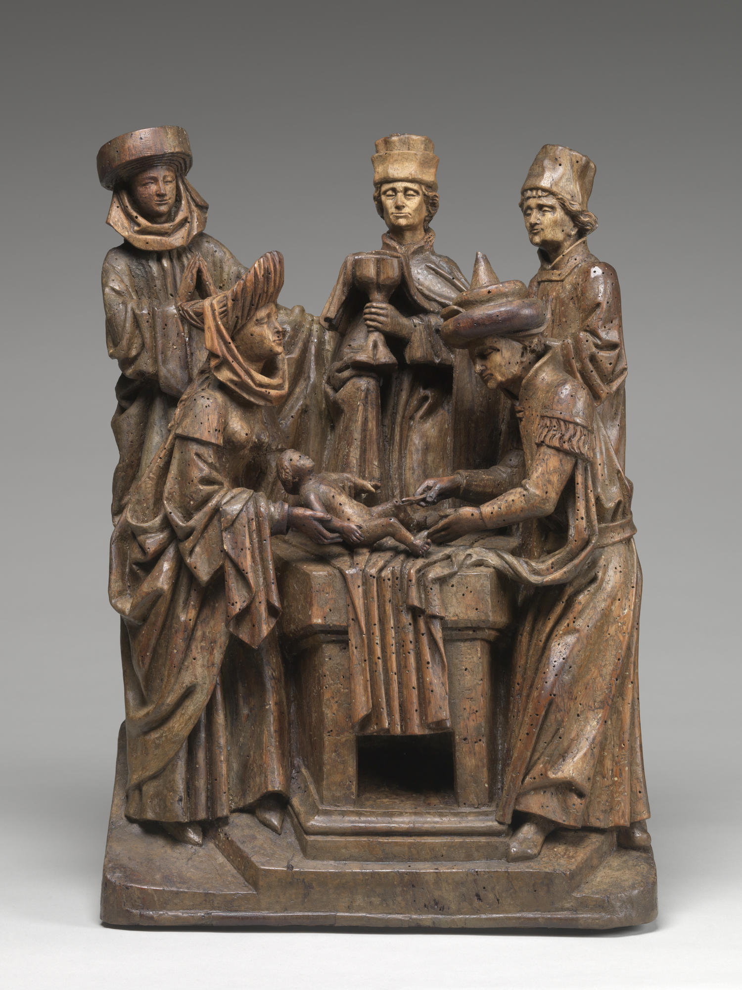 Wood sculpture The Circumcision of Christ, featuring five figures in historic dress standing around a baby lying on an altar.