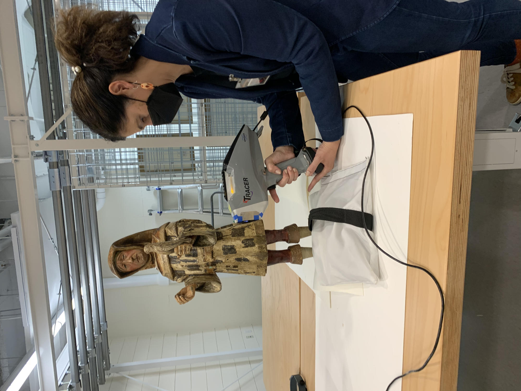  A woman holds a portable XRF instrument along the side of a wood sculpture on a table of a person wearing a tunic and hood.