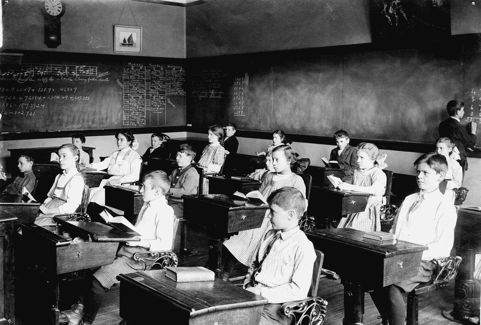 Children at desks in a Cotting School classroom surrounded by blackboards. 