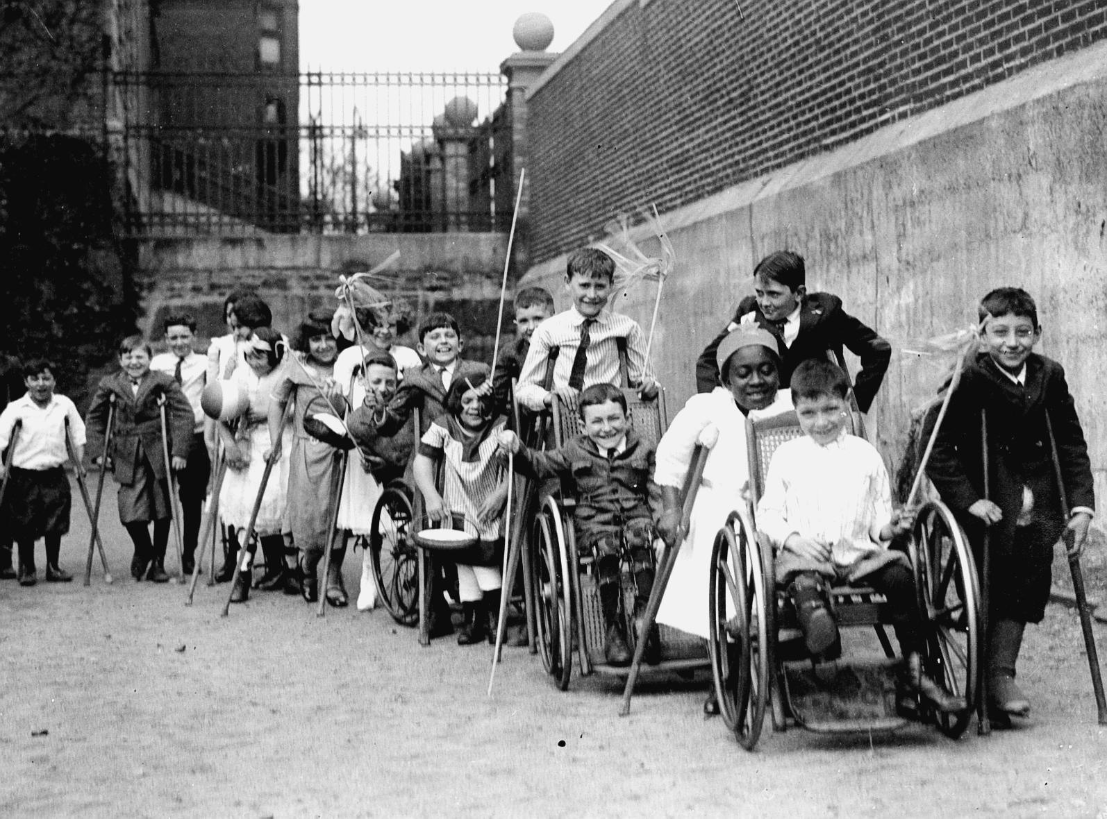 Children who are students at Cotting School, using wheelchairs and crutches at the school lined up single file outdoors. The children are smiling and appear to be having fun.