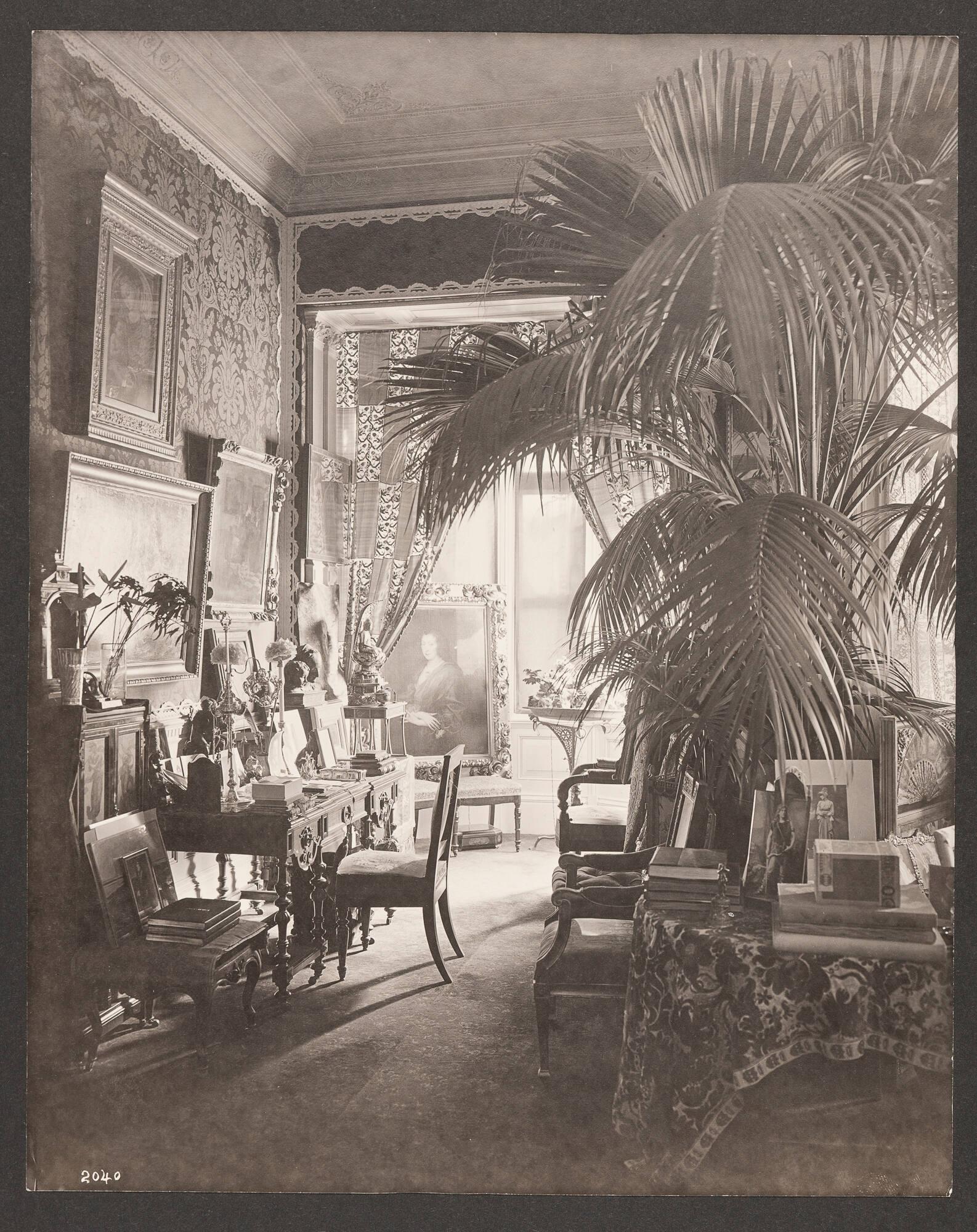 Opulent 19th century domestic interior of Isabella Stewart Gardner’s home crowded with paintings, furniture and decorative arts. The room is dominated by a large potted plant. Paintings line the damask upholstered walls. A round table covered in a velvet floral textile in the foreground is decorated with photographs of musicians and actors and stacks of books.