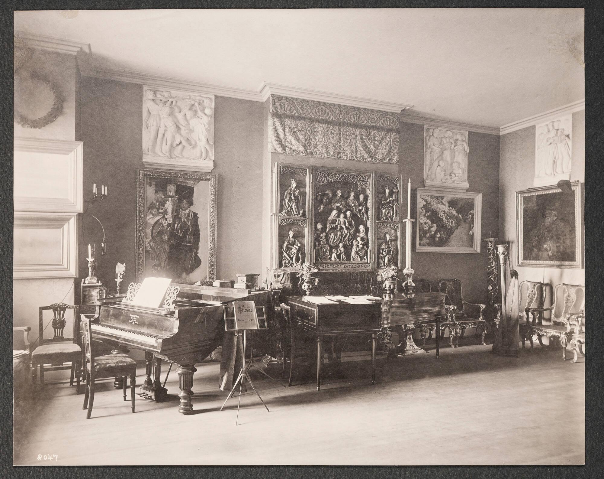 Opulent 19th century domestic interior of Isabella Stewart Gardner’s home, crowded with paintings, furniture and decorative arts. The room contains a piano and a harpsichord. Chairs line the wall hung with paintings and a large wooden altarpiece.  