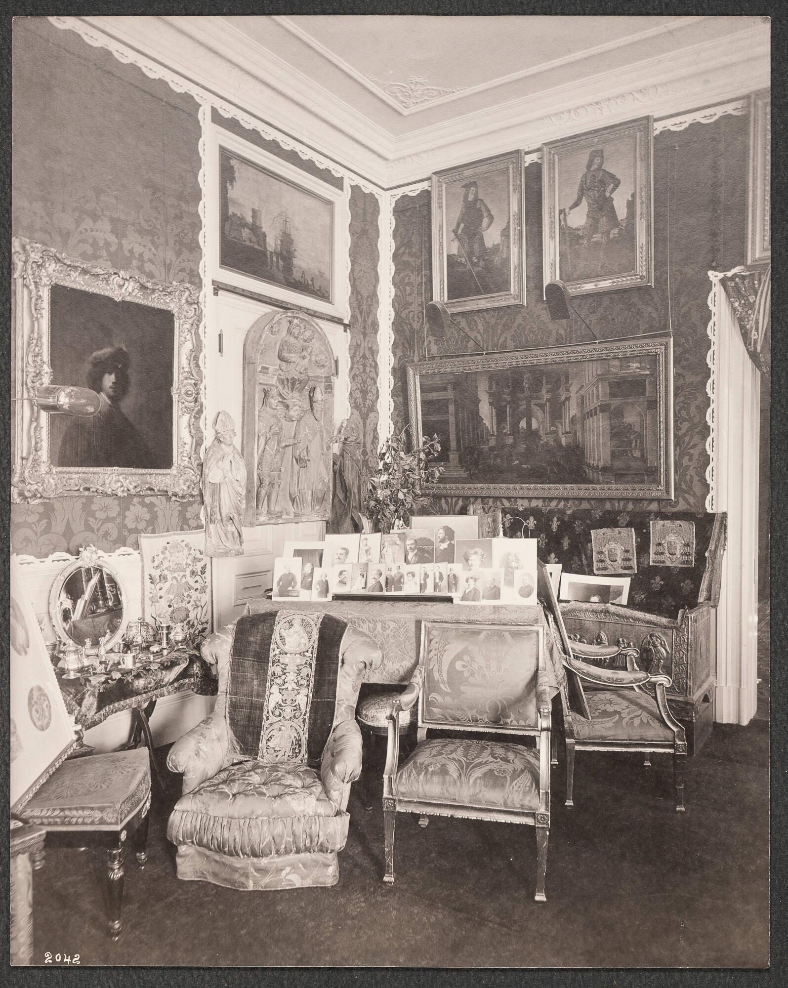 Opulent 19th century domestic interior of Isabella Stewart Gardner’s home crowded with paintings, furniture and decorative arts. Paintings are hung on the damask upholstered walls, chairs surround a table decorated with photographs of musicians. Framed works rest on the seats of empty chairs.