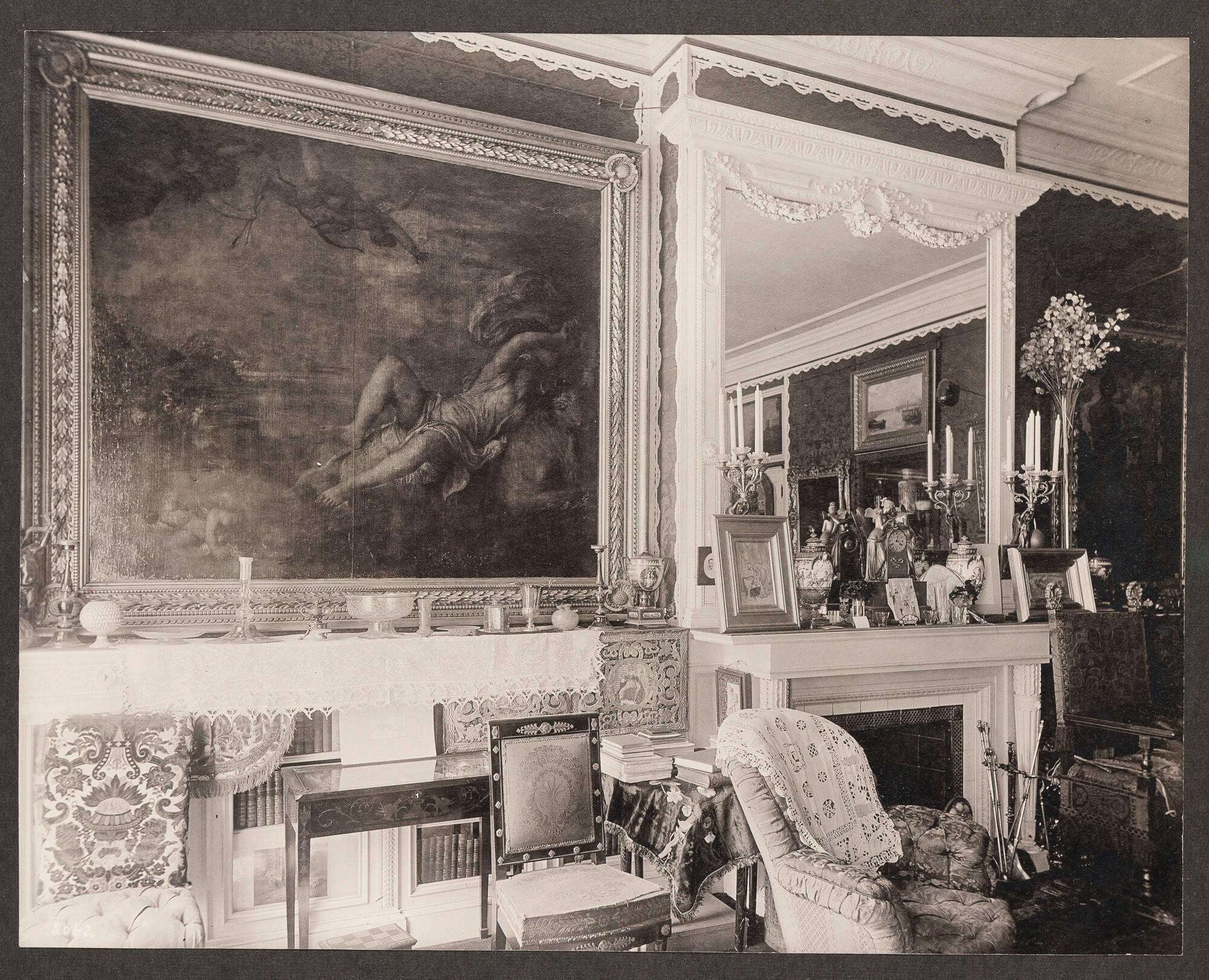 Opulent 19th century domestic interior of Isabella Stewart Gardner’s home crowded with paintings, furniture and decorative arts. Titian’s Europa dominates the foreground. The painting is near a bookcase and a fireplace with a mirror over the mantel. The surfaces are covered with glassware, ceramics, memorabilia and art. An upholstered chair sits in front of the fireplace. 