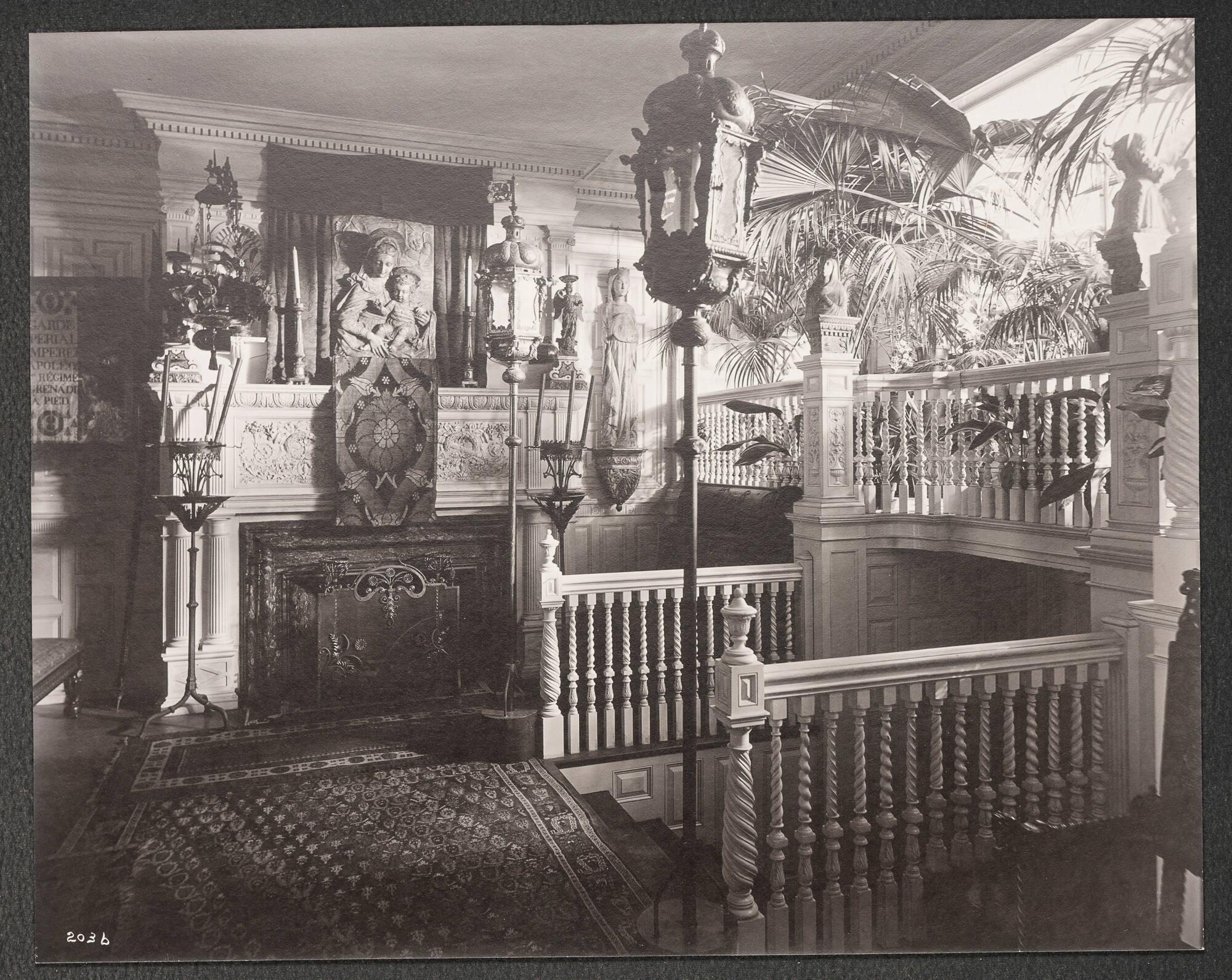 Opulent 19th century domestic interior of Isabella Stewart Gardner’s home, crowded with paintings, furniture, decorative arts. The entryway is dominated by a staircase and balcony bordered by elaborately carved banisters and spiral balustrades. Palm plants and plants reach over from the bay window balcony. 