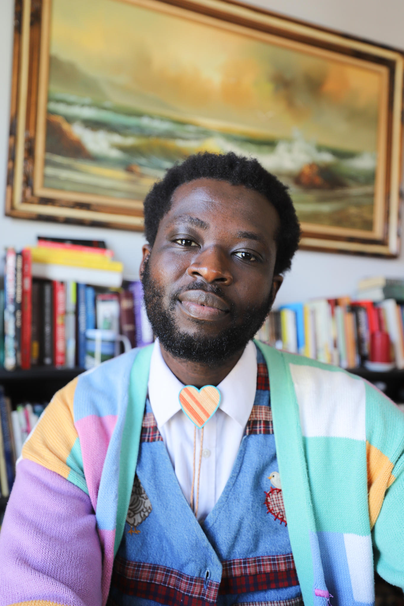 Photo of a person with a painting and bookshelf behind them.