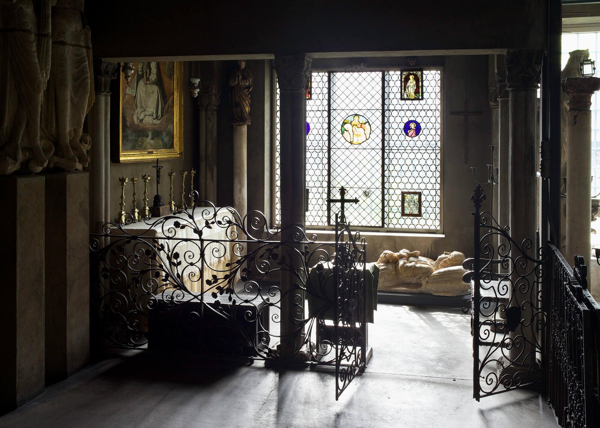 The Spanish Chapel in the Isabella Stewart Gardner Museum, featuring an altar-like installation of a painting of the Virgin Mary and Child above a table draped with a cloth holding a cross and candlesticks is behind a wrought iron gate. Below a window with stained glass is a white marble tomb figure of a Spanish knight.