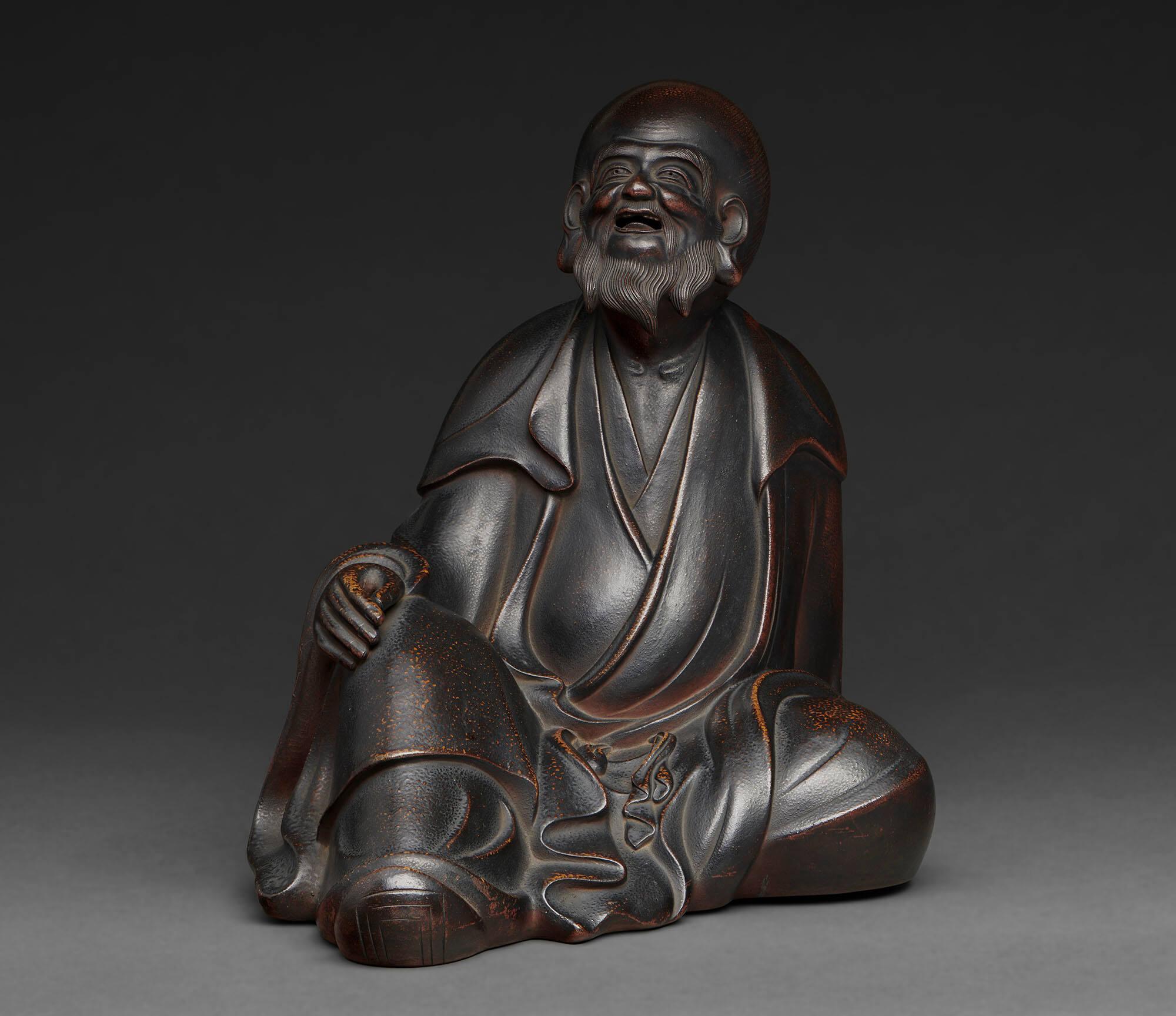 A dark brown to black ceramic figure of a seated man, Chinese poet Li Po. The robed man has a beard and hair pulled into a bun at the back of his head. One hand rests on his knee and the other behind him.
