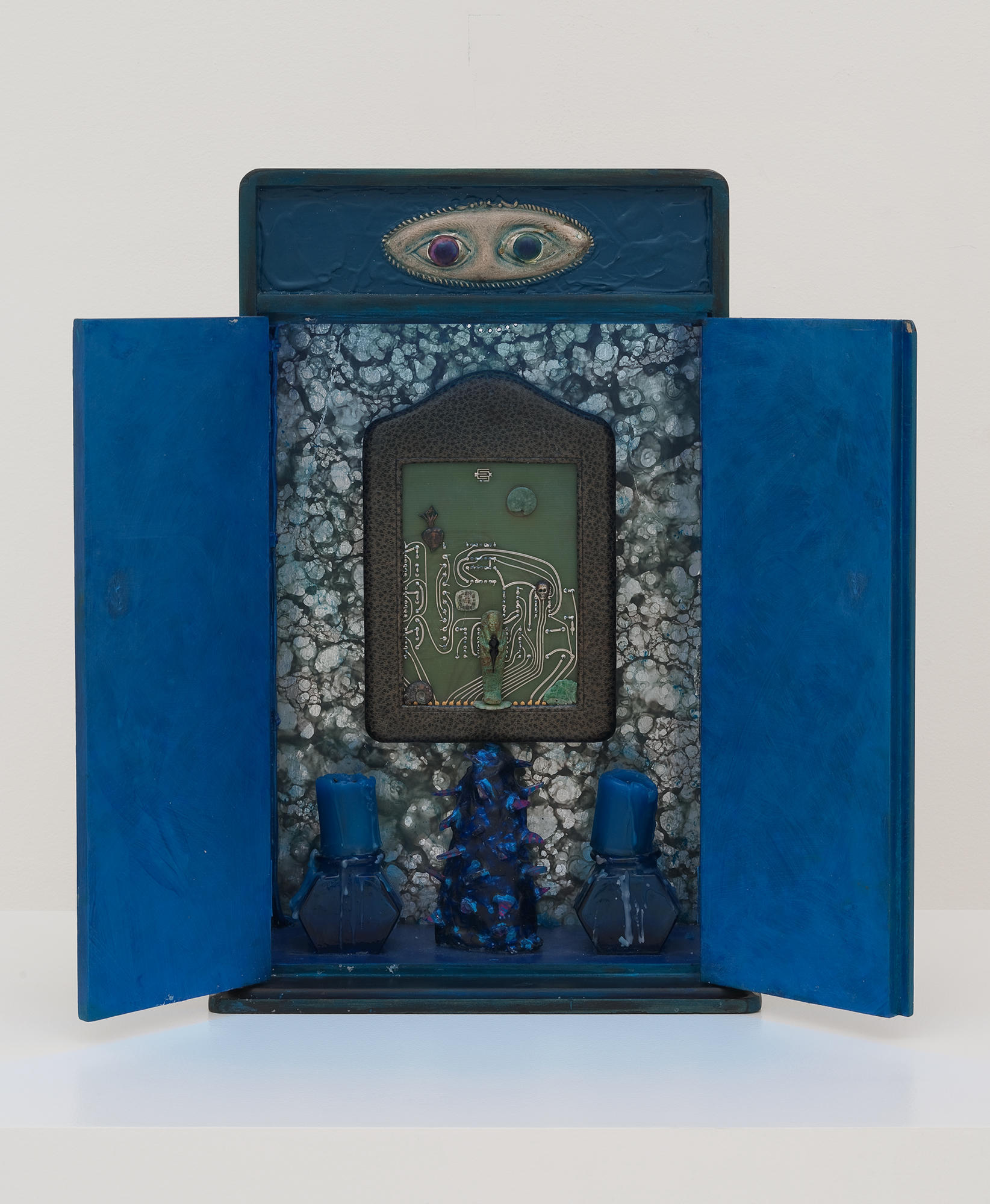 Betye Saar’s Indigo Illusions. An assembled altar-like sculpture in tones of blue has a luggage tag with a drawing above two burned down blue candles. Two open eyes are on the top of the work.