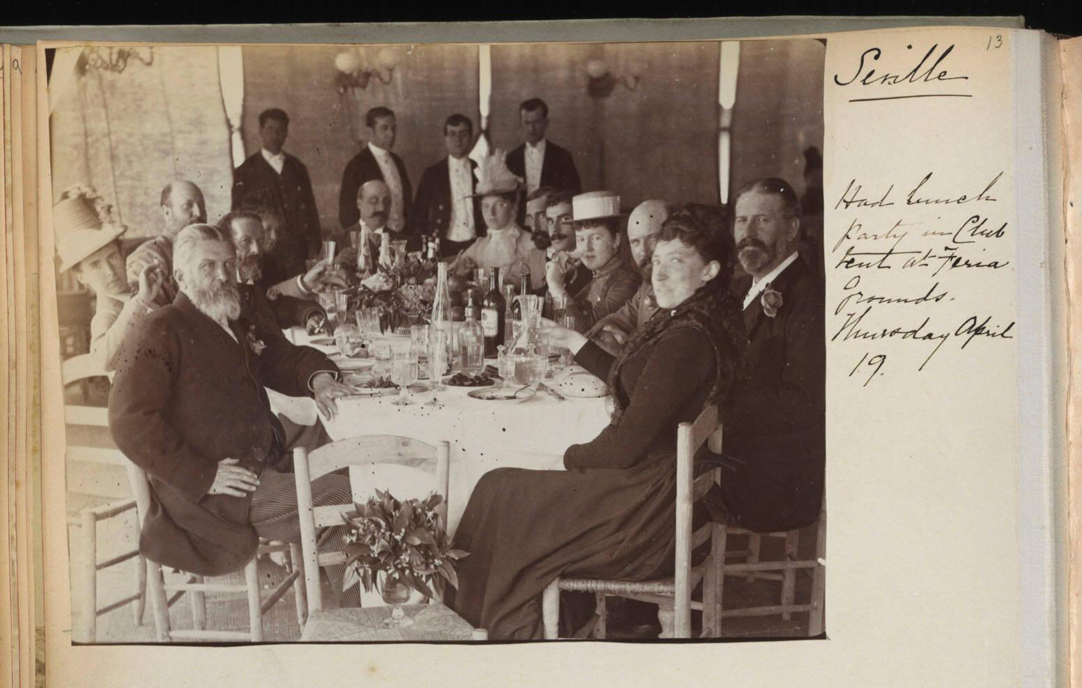 The top half of a page from a travel album with a black and white photograph of thirteen people around a dining table. The page is annotated in ink in Isabella Stewart Gardner’s hand “Seville, had lunch in Club Tent at Feria Grounds. Thursday April 19.”