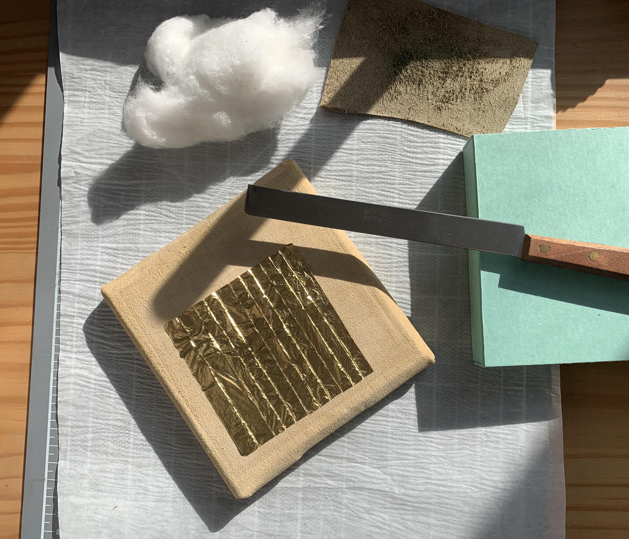 A thin sheet of genuine 22 karat gold cut into long strands laid over a soft leather covered square pillow with a gold knife and clean cotton nearby.