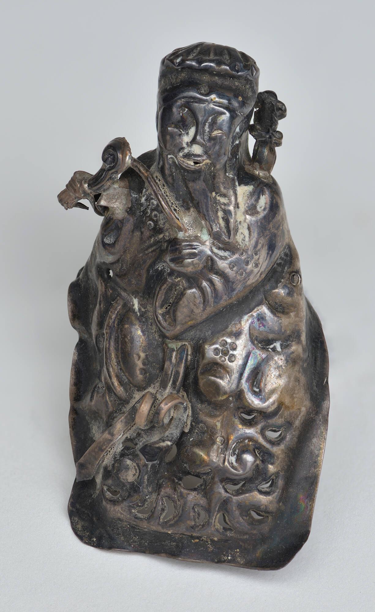 A tarnished silver object with a high relief design of a figure wearing a robe and headdress shown before conservation at the Isabella Stewart Gardner Museum.