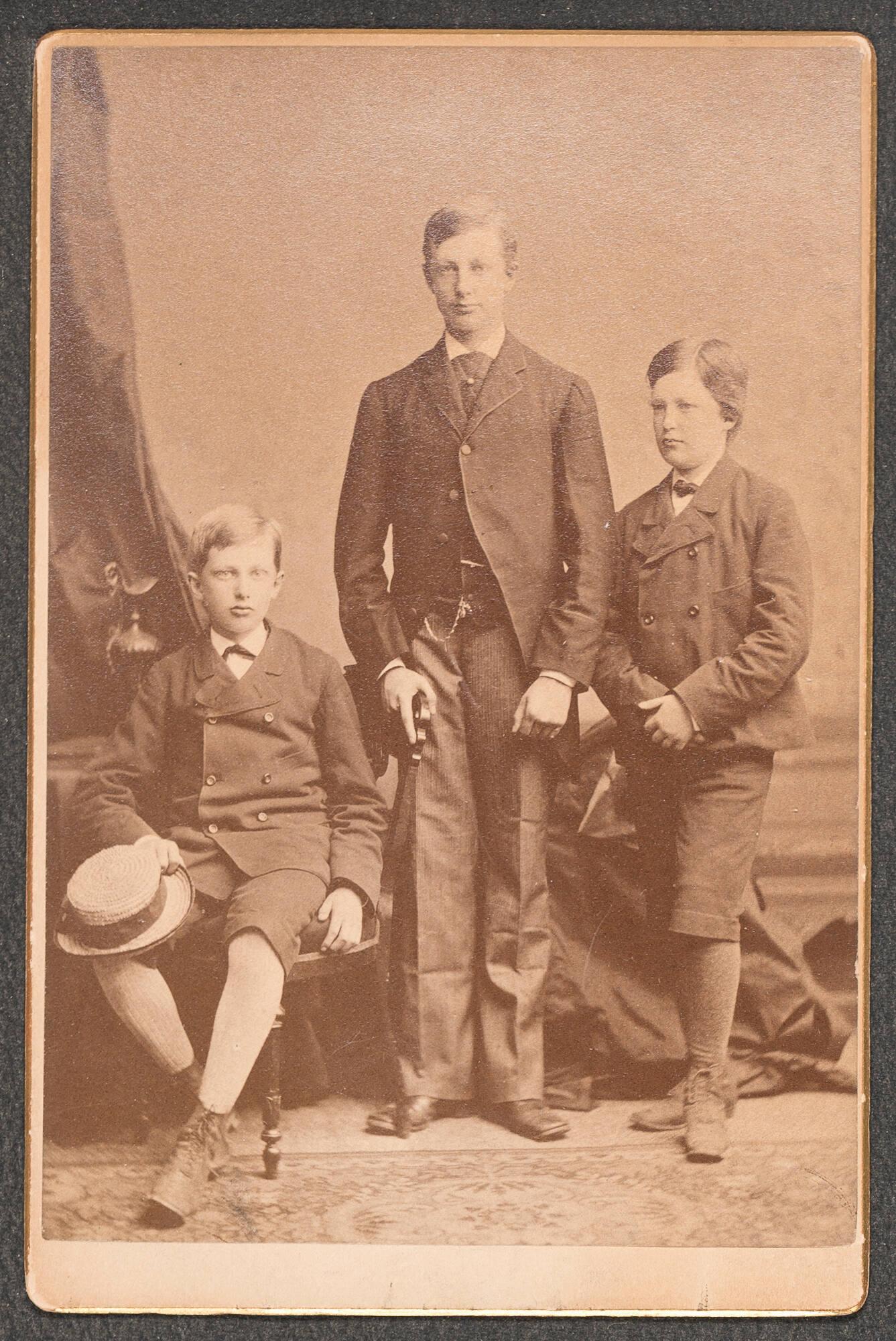 A sepia toned photographed of three white boys in 19th century dress. The oldest teenage boy, Joseph Peabody Gardner, is in a dark suit with long pants. The younger boys, William Amory Gardner, 12, and August Peabody Gardner, 10, are in dark jackets and short pants.