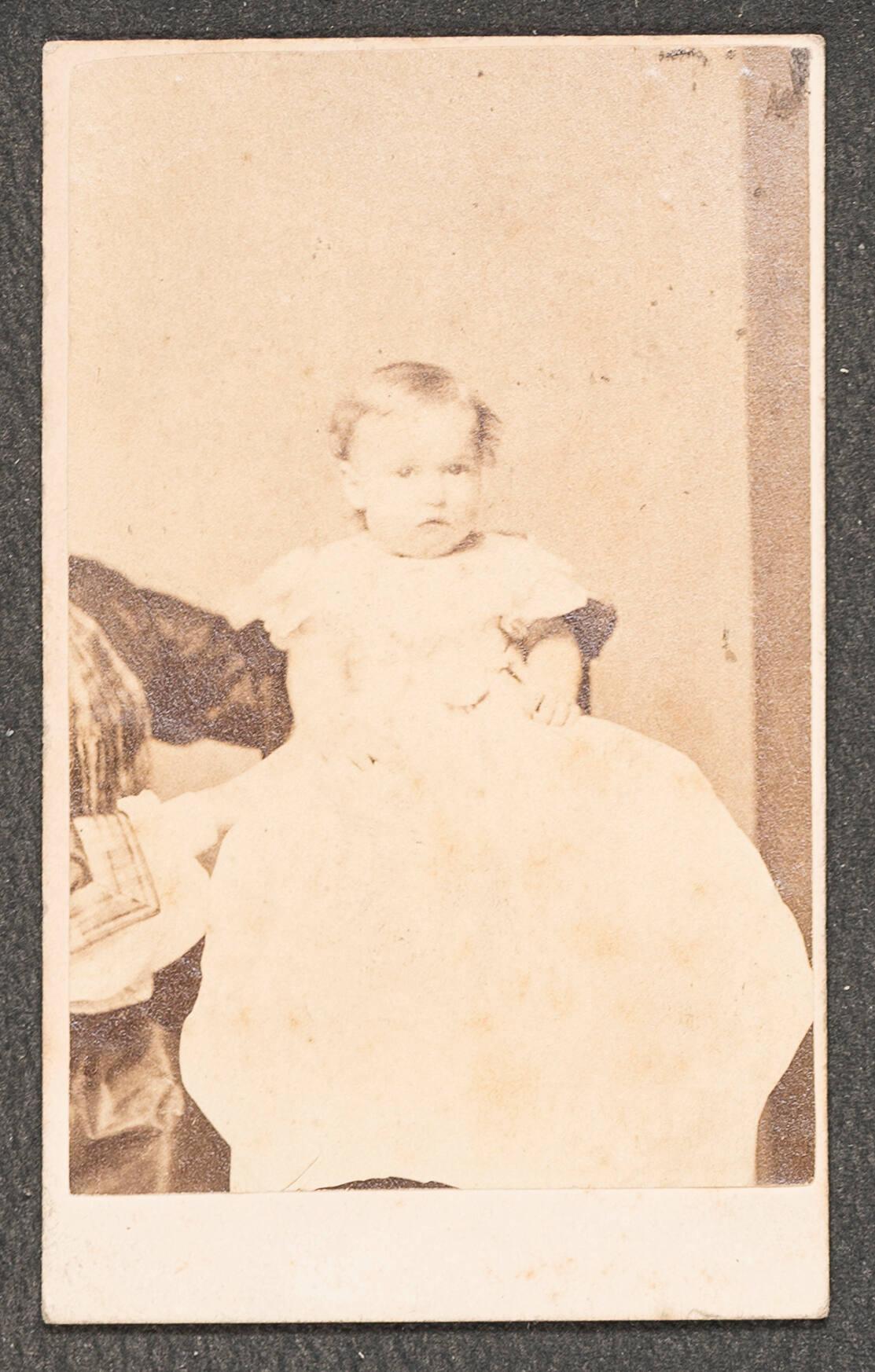 A sepia toned photograph of a 9 month old white baby in a white gown. The infant, Jackie Gardner, is seated and being supported by the arms of his mother, Isabella Stewart Gardner, who is mostly off camera.