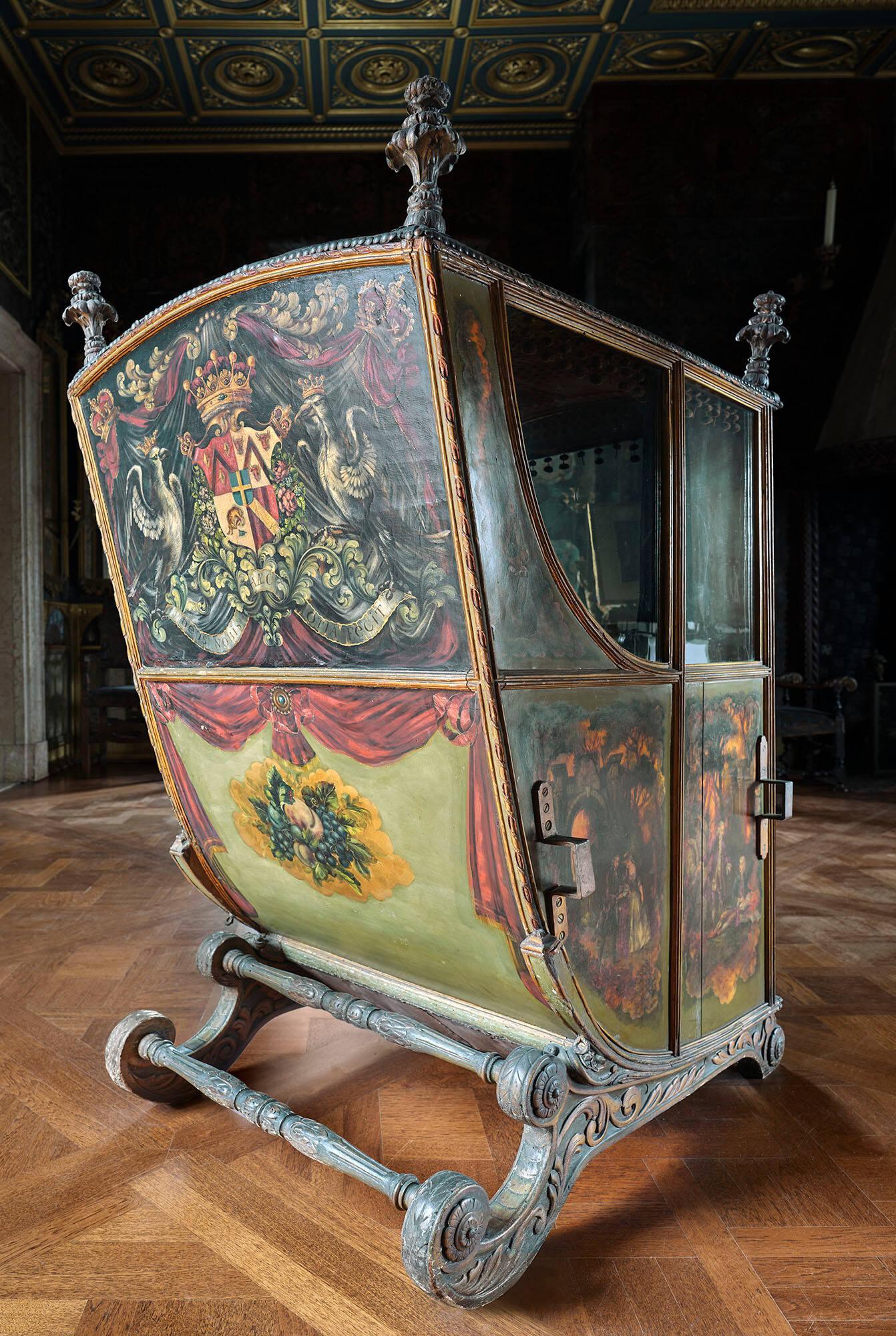 Back of the sedan chair in the Gardner Museum’s Veronese Room, with a large painting featuring a coat of arms and two crown-wearing birds surrounded by sprawling greenery and holding a ribbon adorned with text