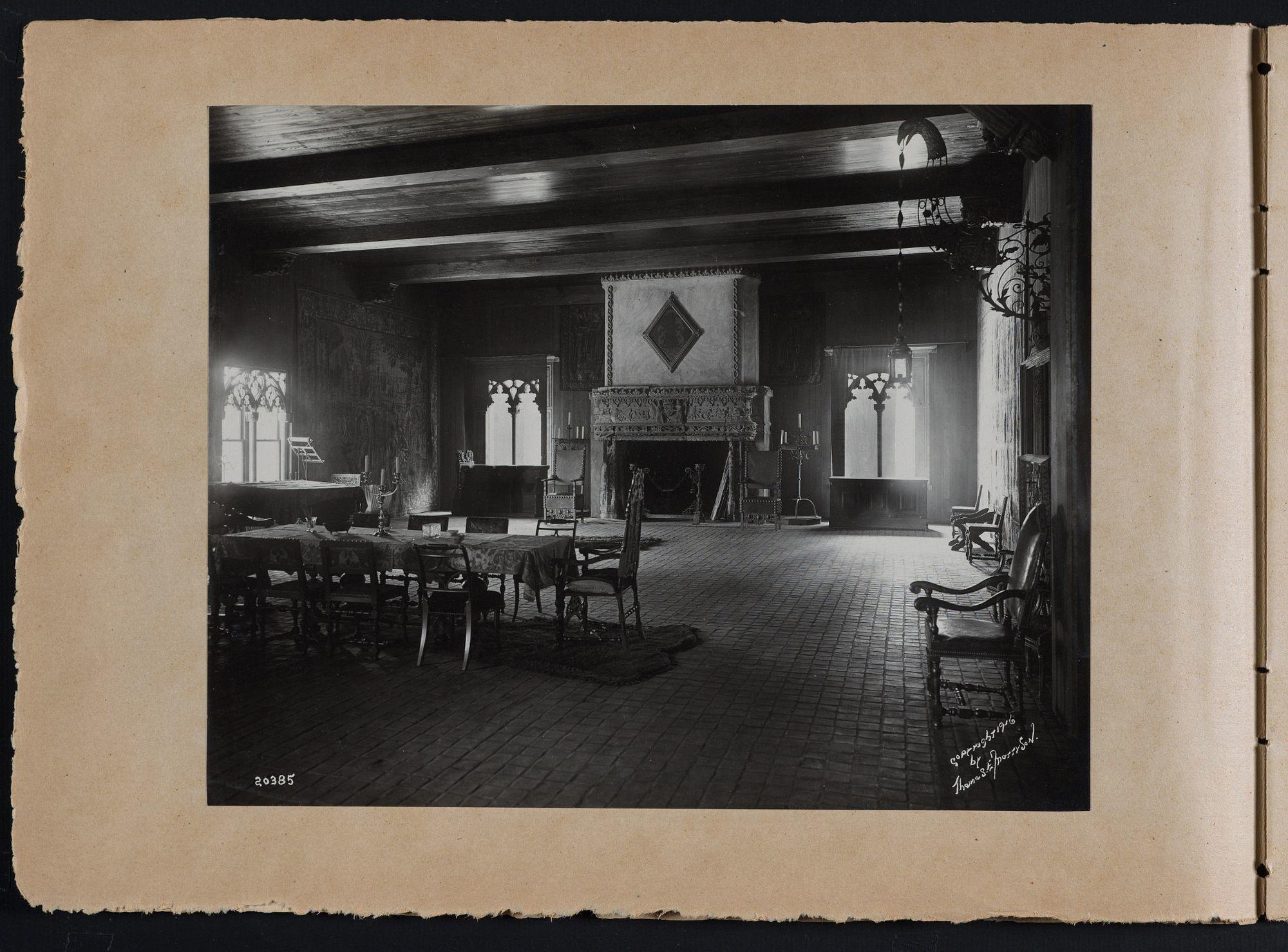 The Tapestry Room at the Isabella Gardner Museum. A table in the foreground is set with dishes and surrounded by chairs. A large carved stone fireplace is visible in the background. 