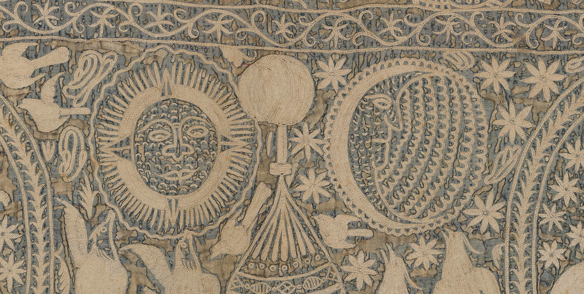 A detail of an embroidered wall hanging in the Gardner Museum of cream colored thread on a dark blue background. On either side of a spire is a sun and a moon, both with faces.