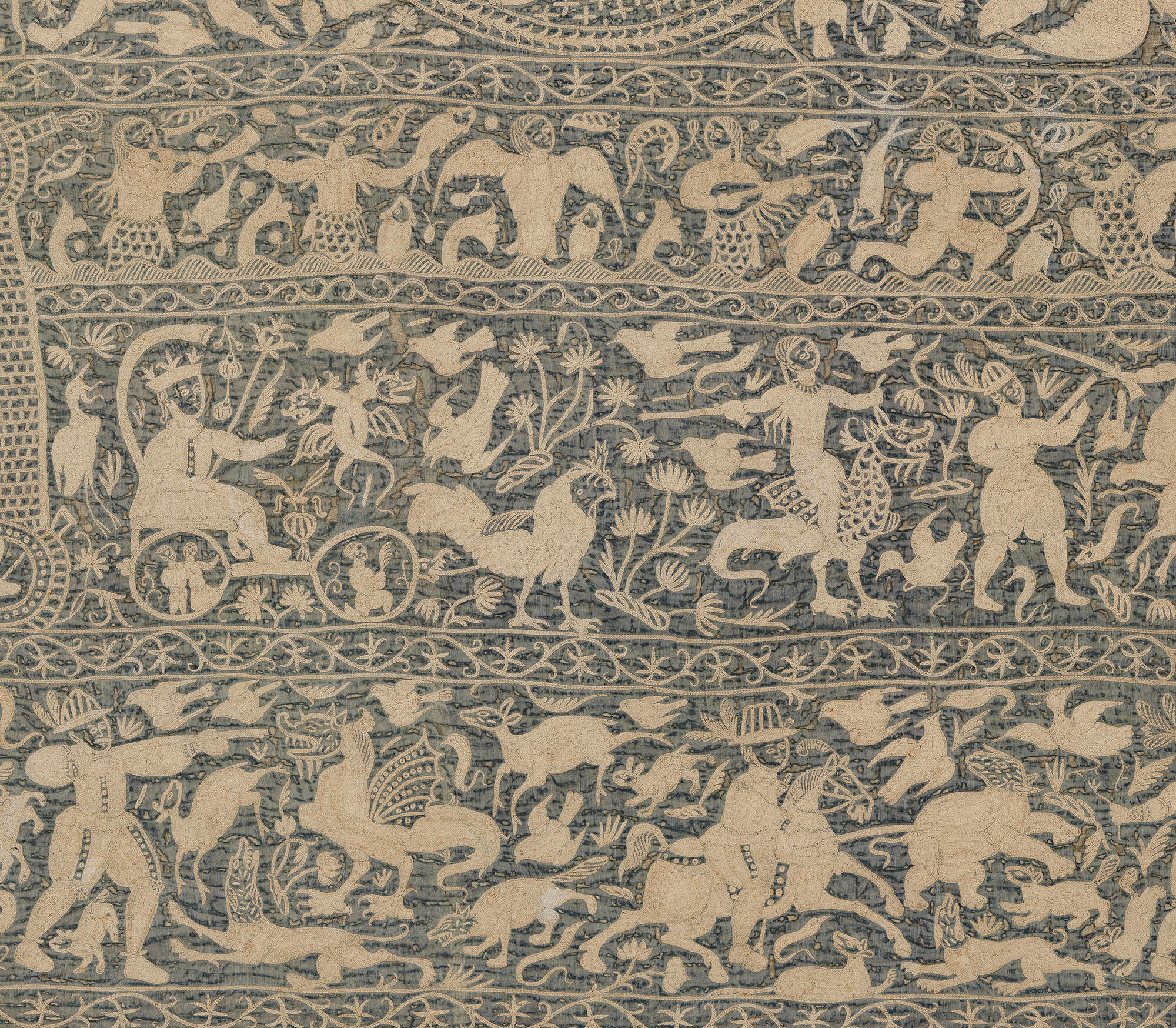 A detail of an embroidered wall hanging in the Gardner Museum of cream colored thread on a dark blue background. A crowned figure is in a chariot pulled by a rooster and surrounded by mythological creatures.