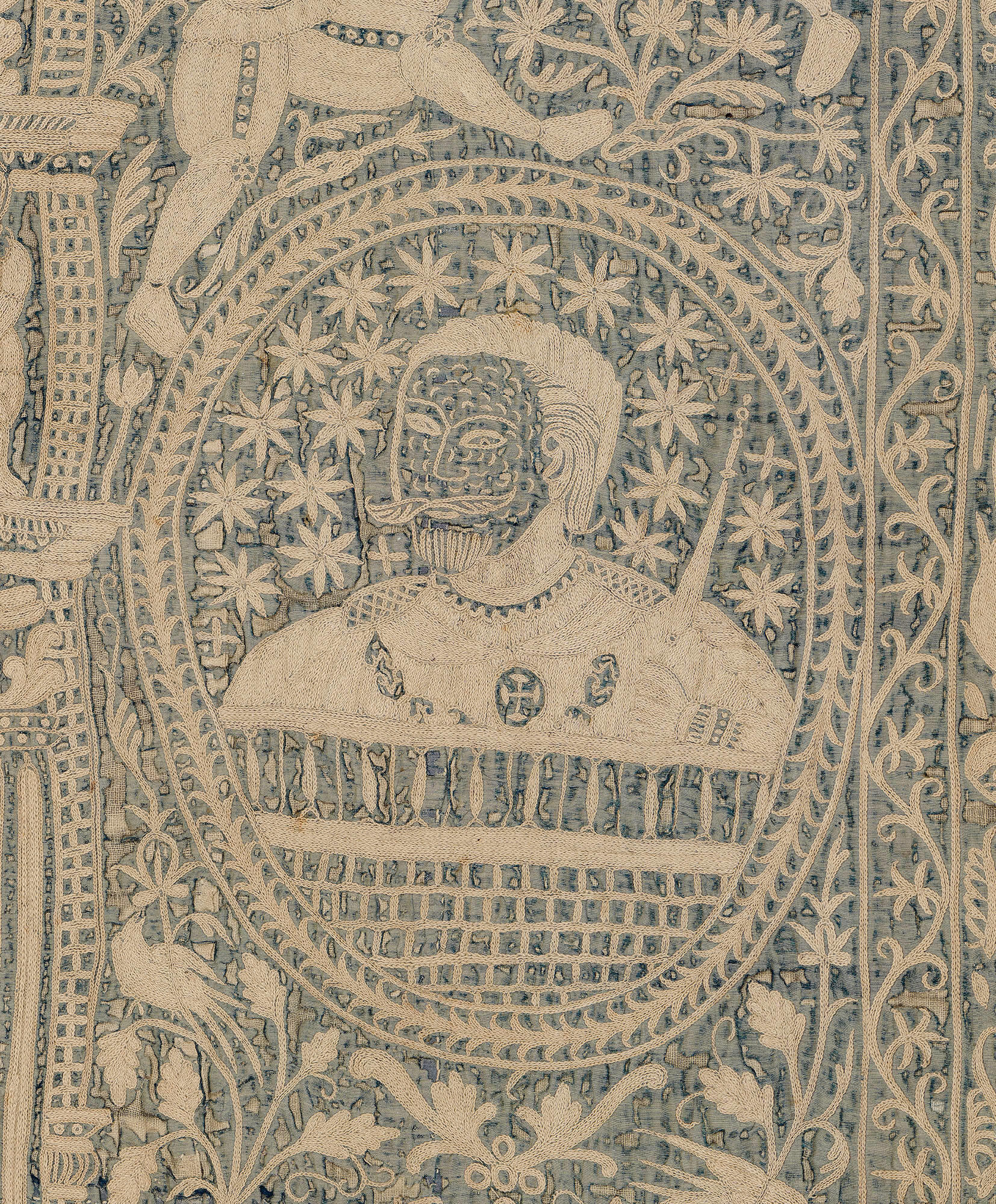 A detail of an embroidered wall hanging in the Gardner Museum of cream colored thread on a dark blue background. The bust of a man with short hair and mustache is in an oval frame.