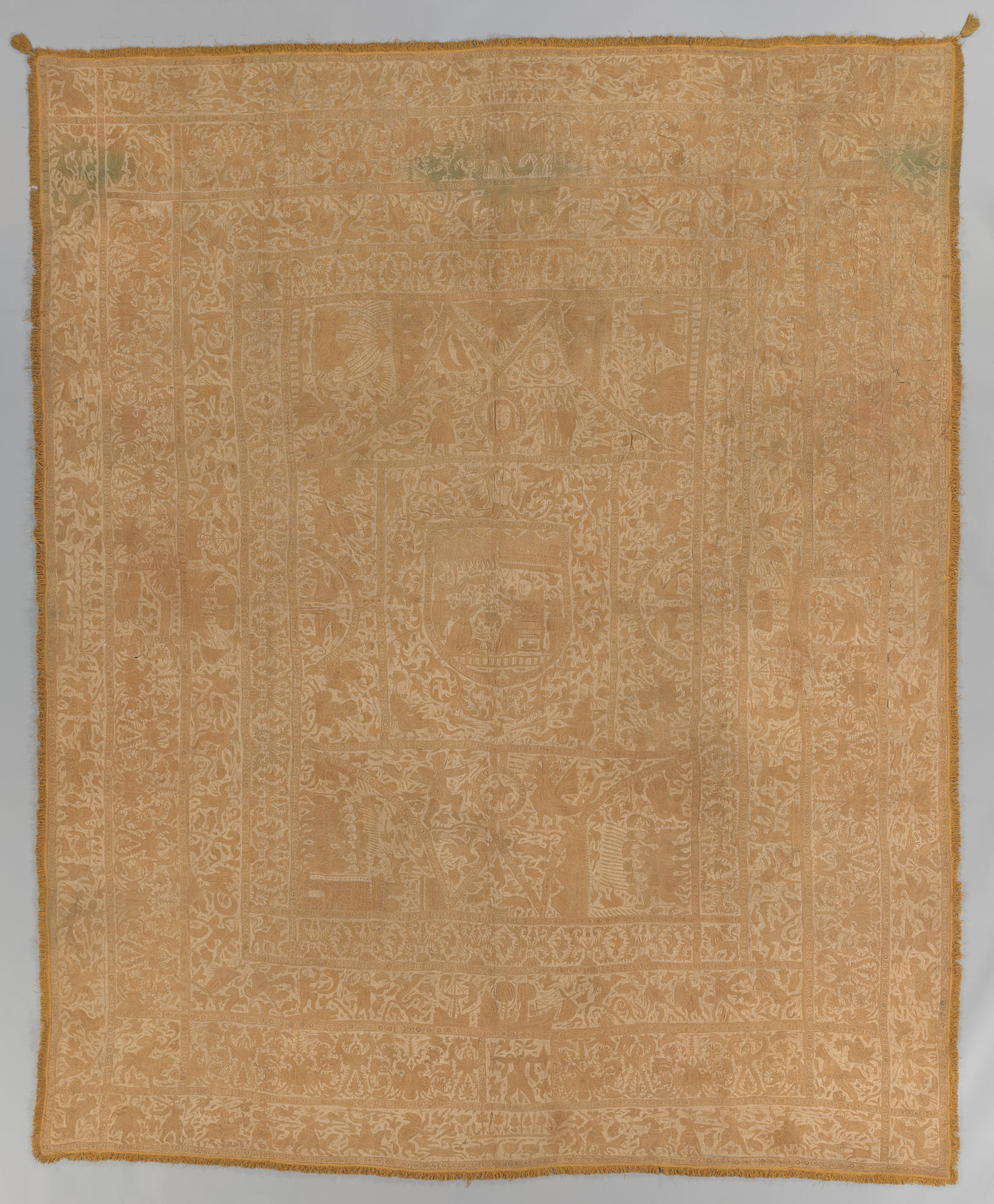An embroidered colcha, or bed cover, of cream colored thread on a mustard yellow background. Four rows of borders with people, plants, animals, and mythological creatures surround a central field a king at court