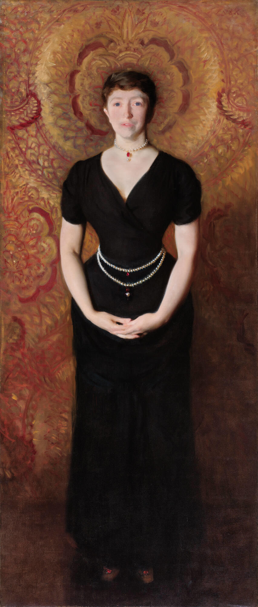 A woman, Isabella Stewart Gardner, with light skin, brown hair, and blue eyes stands with her hands clasped in front of her. She wears a black dress with pearls and rubies around her neck and waist.